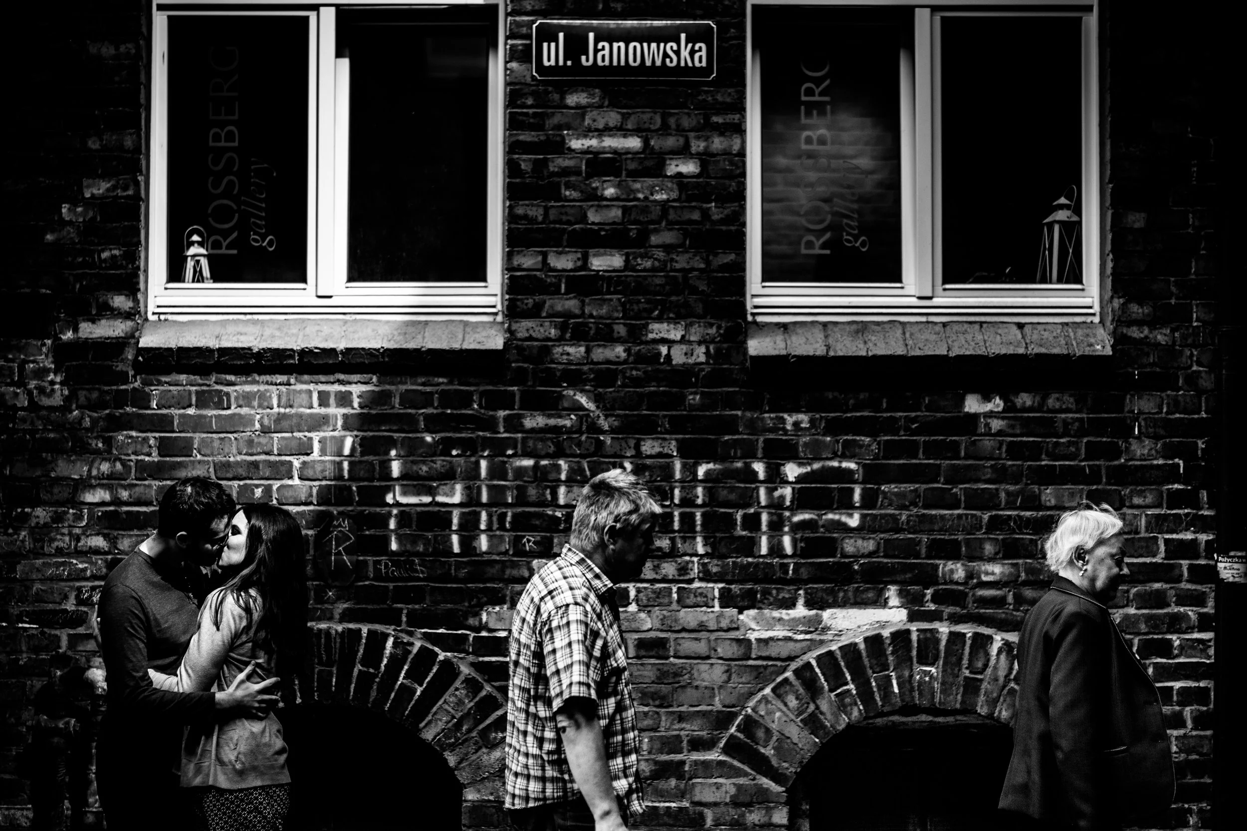 Black and white photo of a brick building with a street sign 'ul. Janowska' and three people walking past, two embracing and kissing, one walking alone, with windows displaying 'RоскопBERG gallery' and small lanterns inside.
