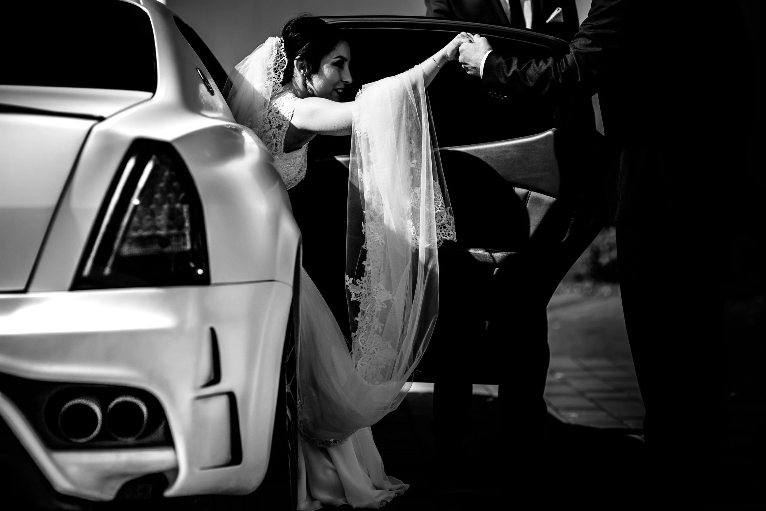 A bride in a wedding dress and veil is sitting in a car, smiling, as she reaches out to hold hands with someone outside the car.