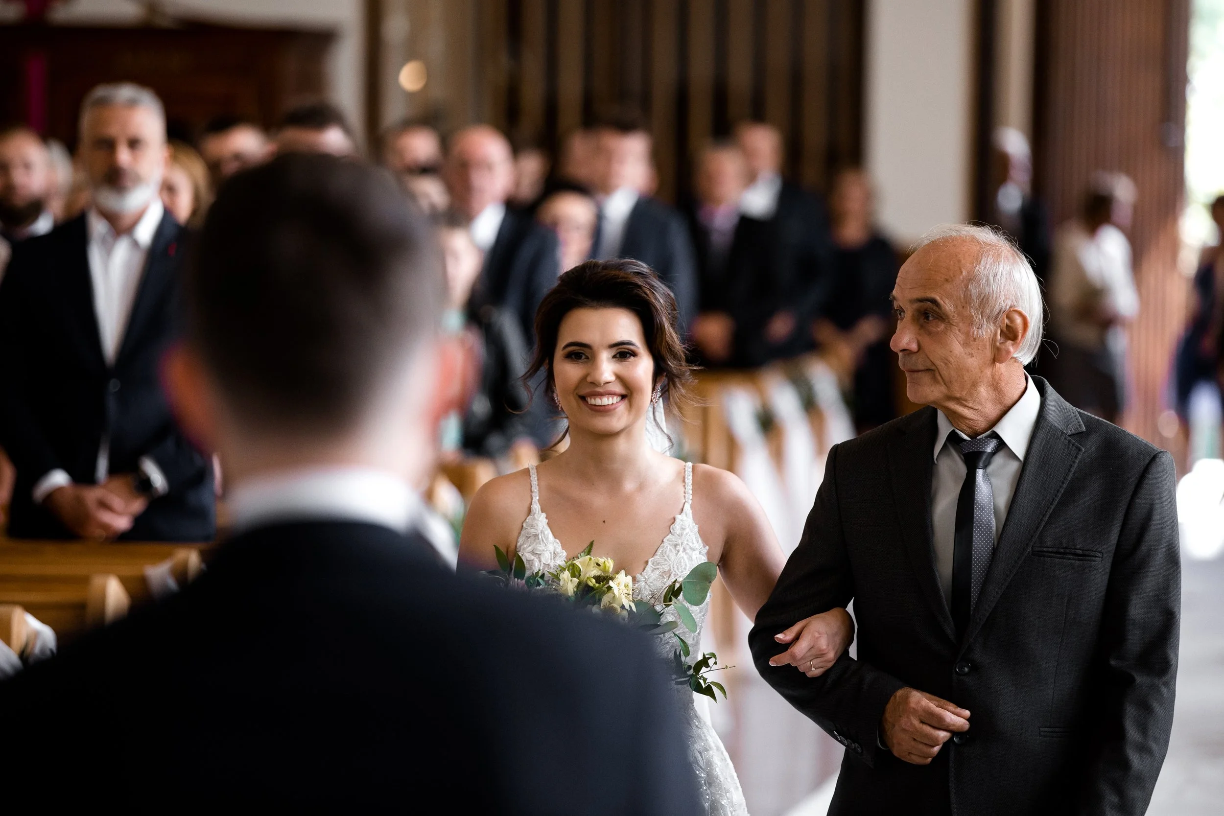 Uśmiechnięta panna młoda idzie do ołtarza ze swoim ojcem podczas ceremonii ślubnej, w tle widać gości.