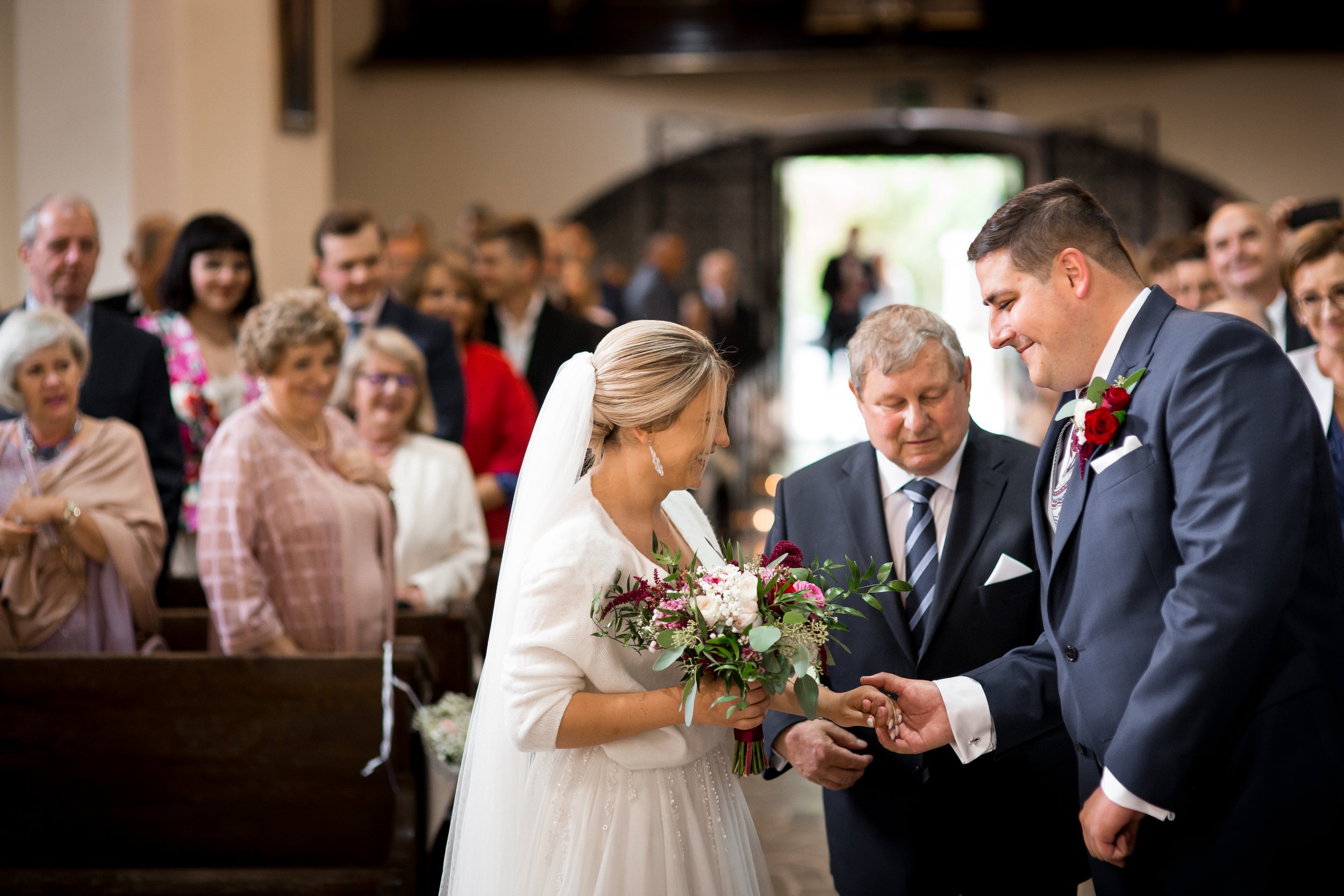 A bride and groom exchanging rings in front of a priest during a wedding ceremony in a church, with guests smiling and watching in the background.