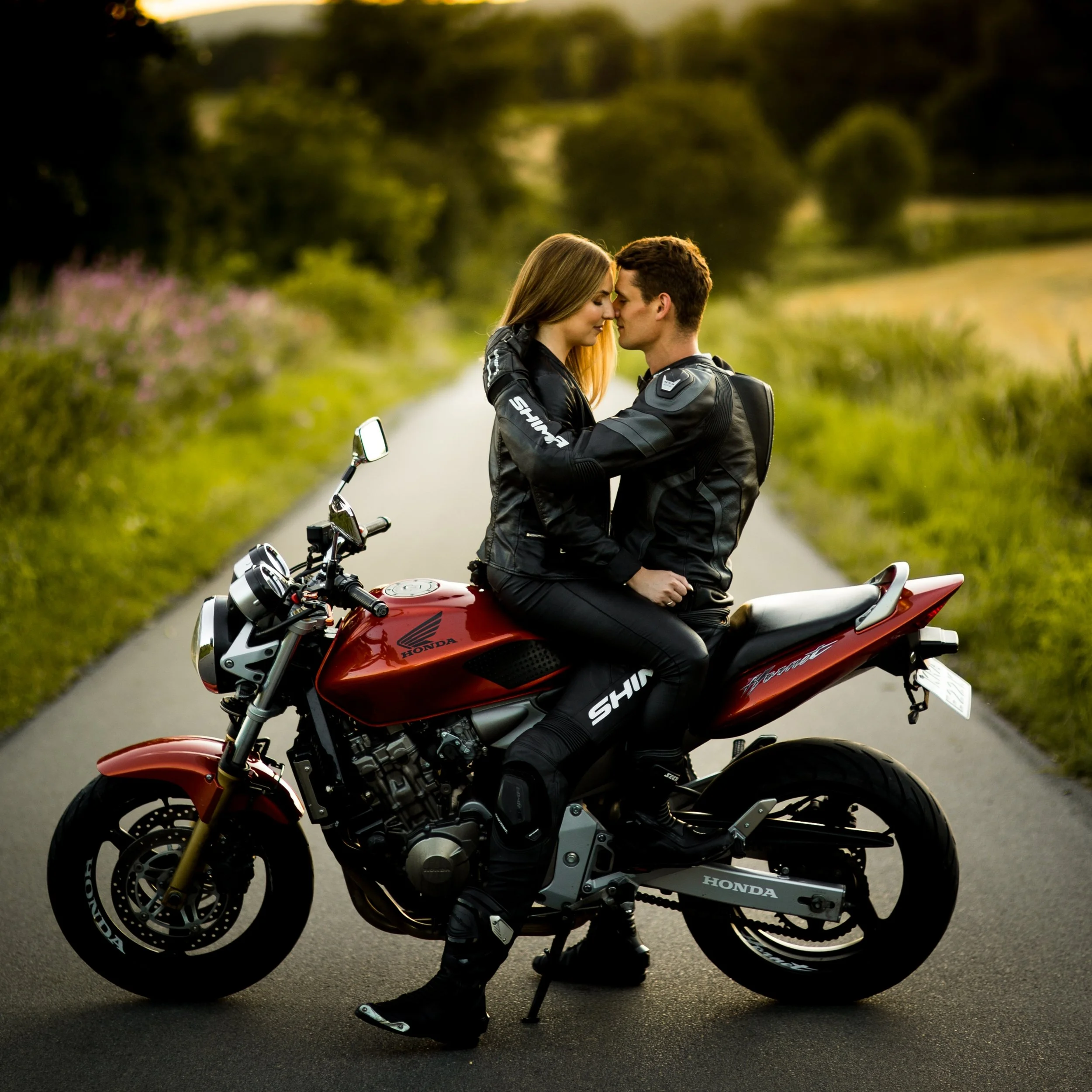 A couple in motorcycle gear sitting on a red Honda motorcycle on a rural road, embracing each other with nature in the background.
