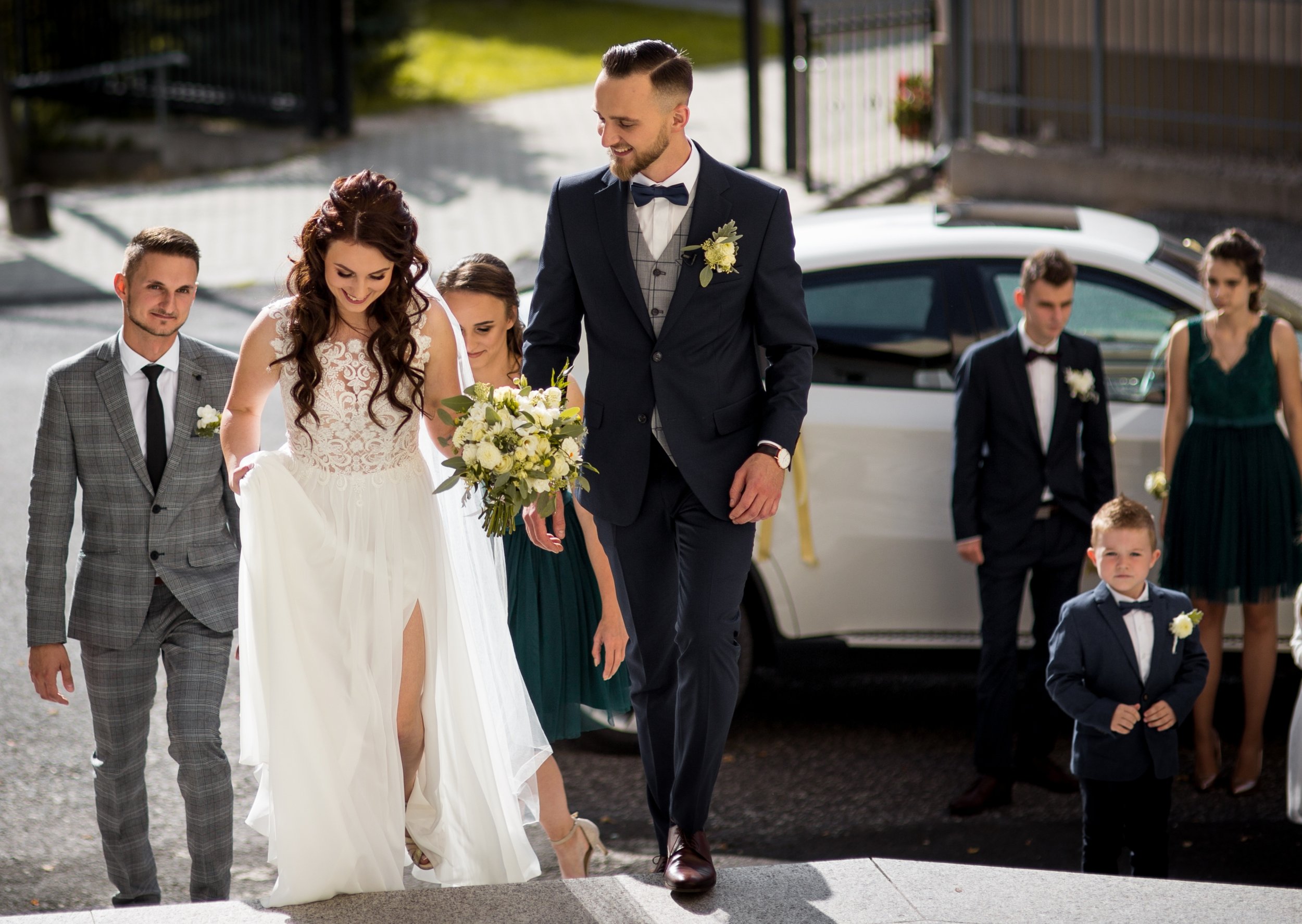 A bride and groom walking down steps, surrounded by wedding party members, with a white car in the background.