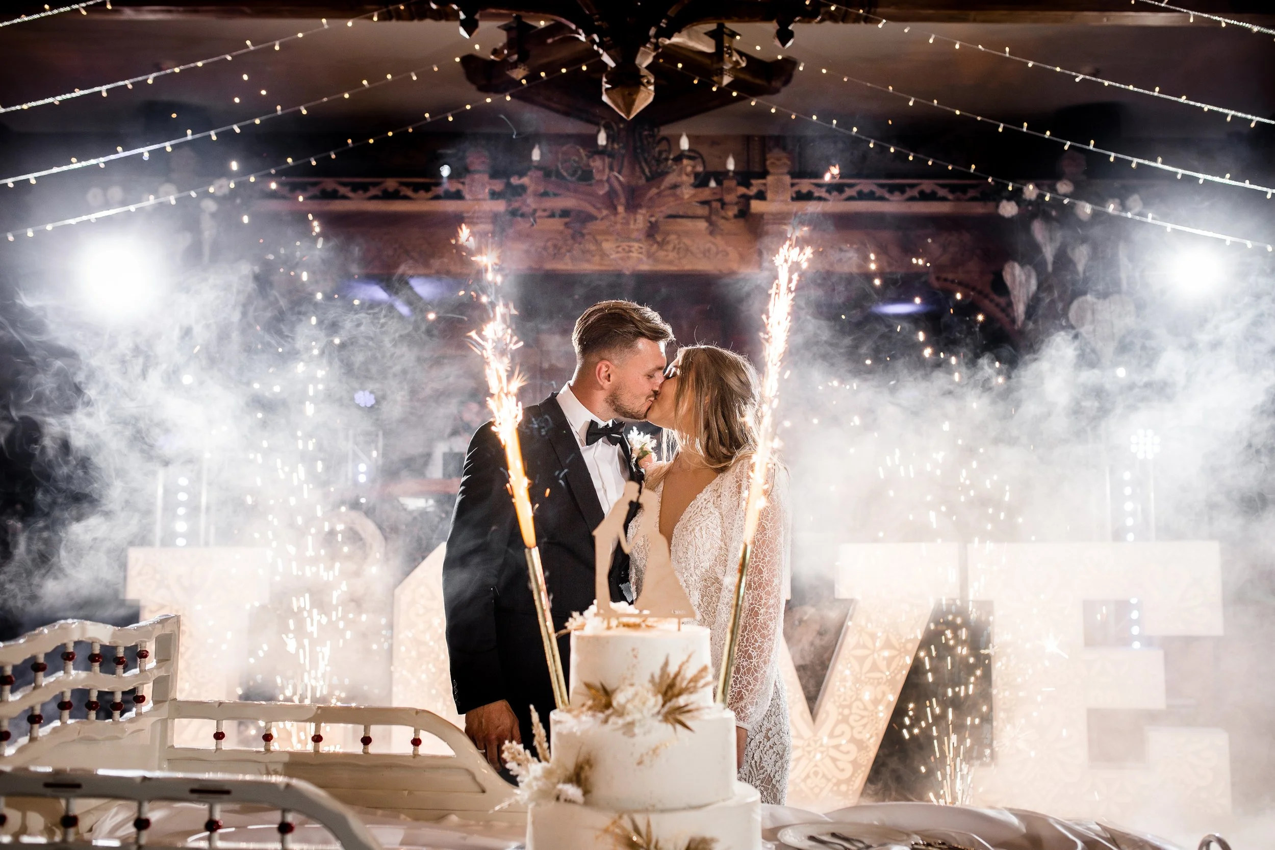 Para młoda całuje się podczas przyjęcia weselnego, na pierwszym planie widać zimne ognie i tort weselny.