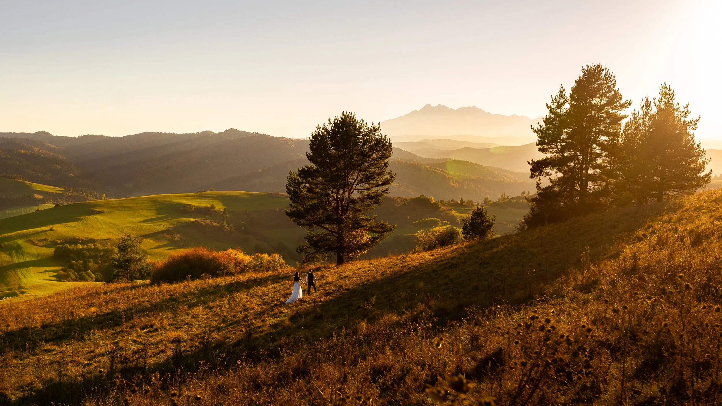 Para spaceruje trzymając się za ręce po trawiastym zboczu wzgórza o zachodzie słońca. W tle widać drzewa, pagórki i odległe góry.