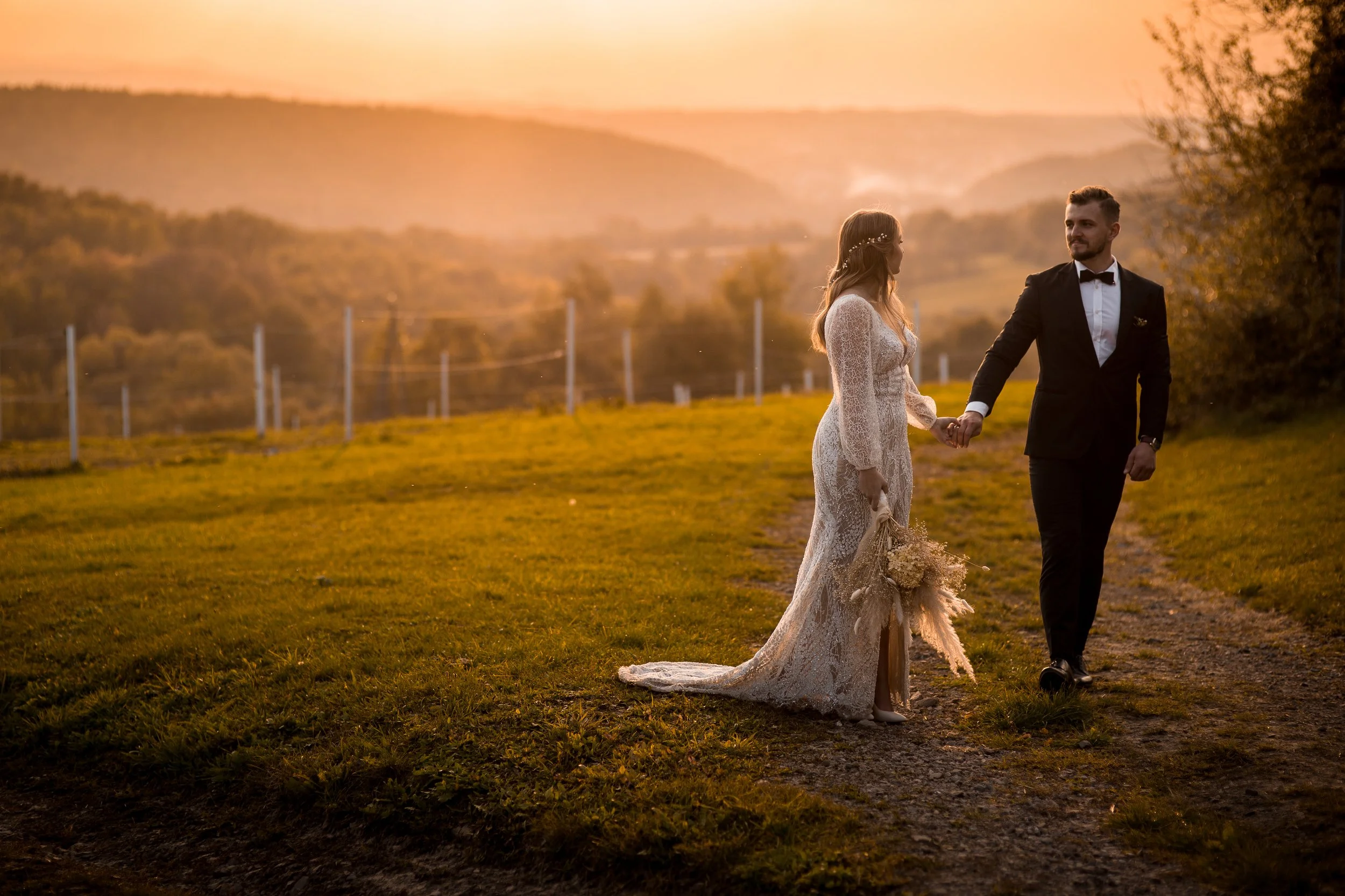 A bride and groom holding hands on a dirt path at sunset in a scenic rural landscape