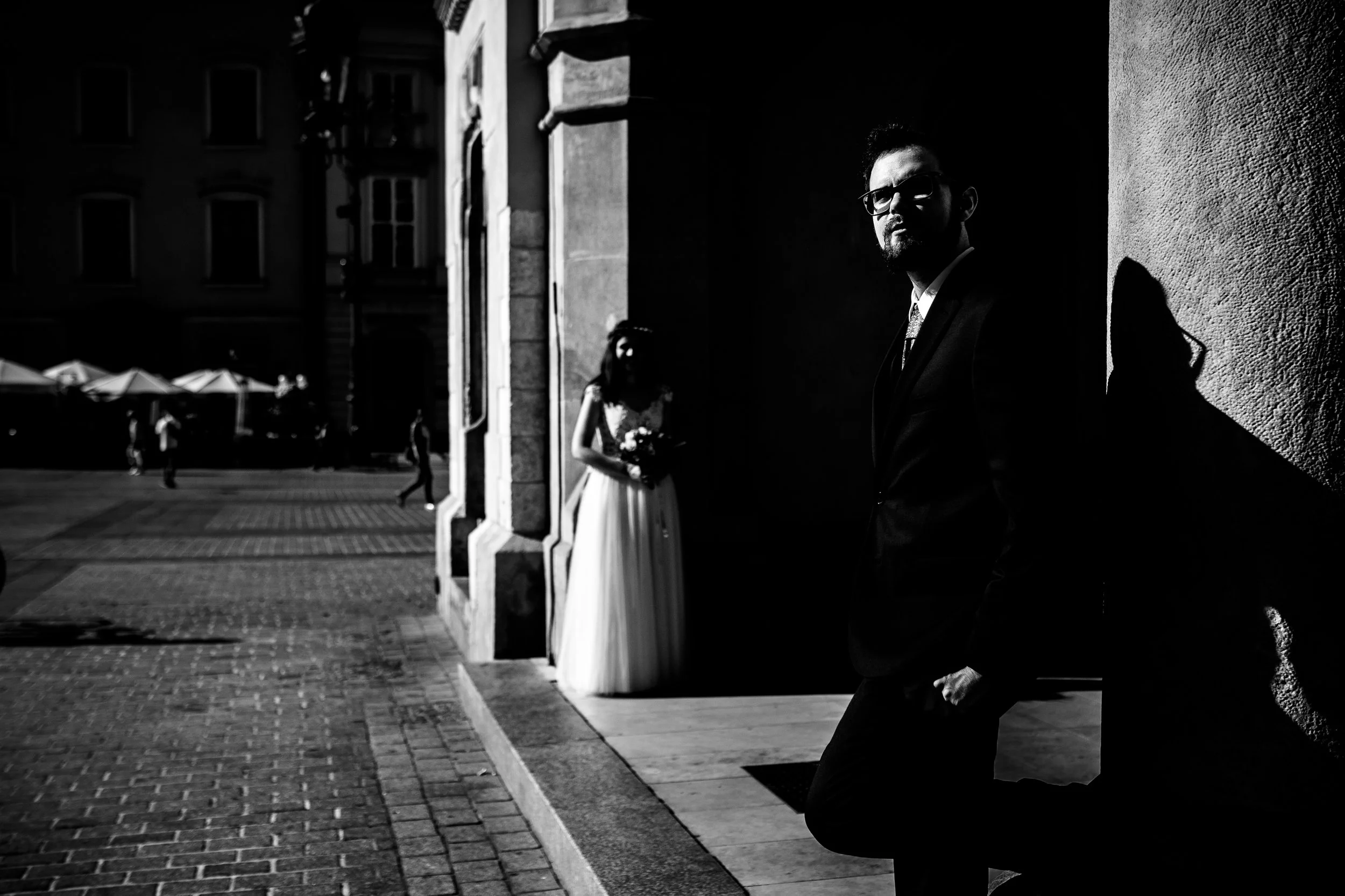 A man in a suit and glasses leaning against a wall in shadow, with a bride in a wedding dress holding a bouquet in the bright background outside on a city street.
