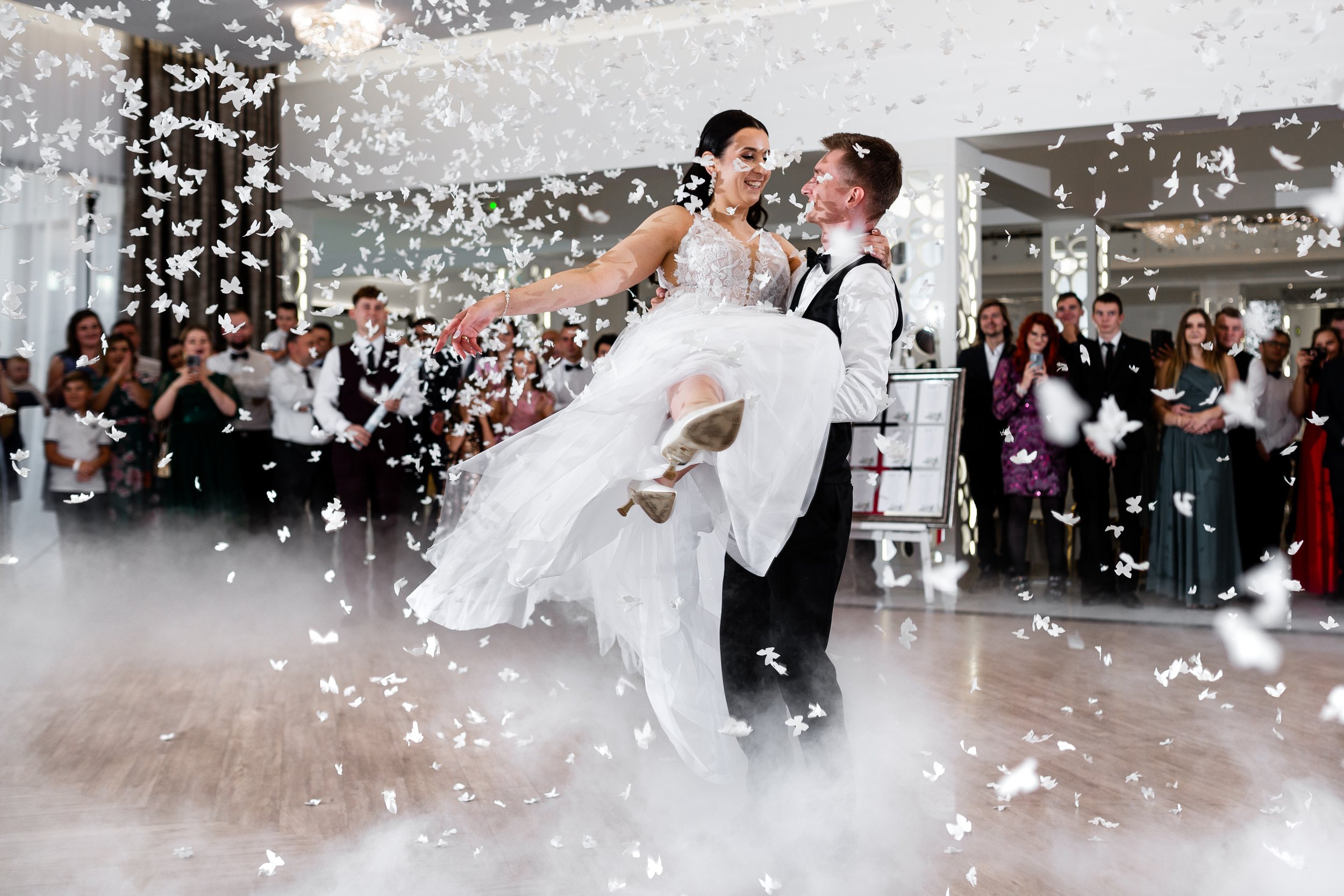 Młoda para tańczy na przyjęciu weselnym, pan młody trzyma pannę młodą, która unosi rękę, a wokół niego kręcą się członkowie rodziny i przyjaciele. Na parkiecie widać konfetti i efekty dymu.