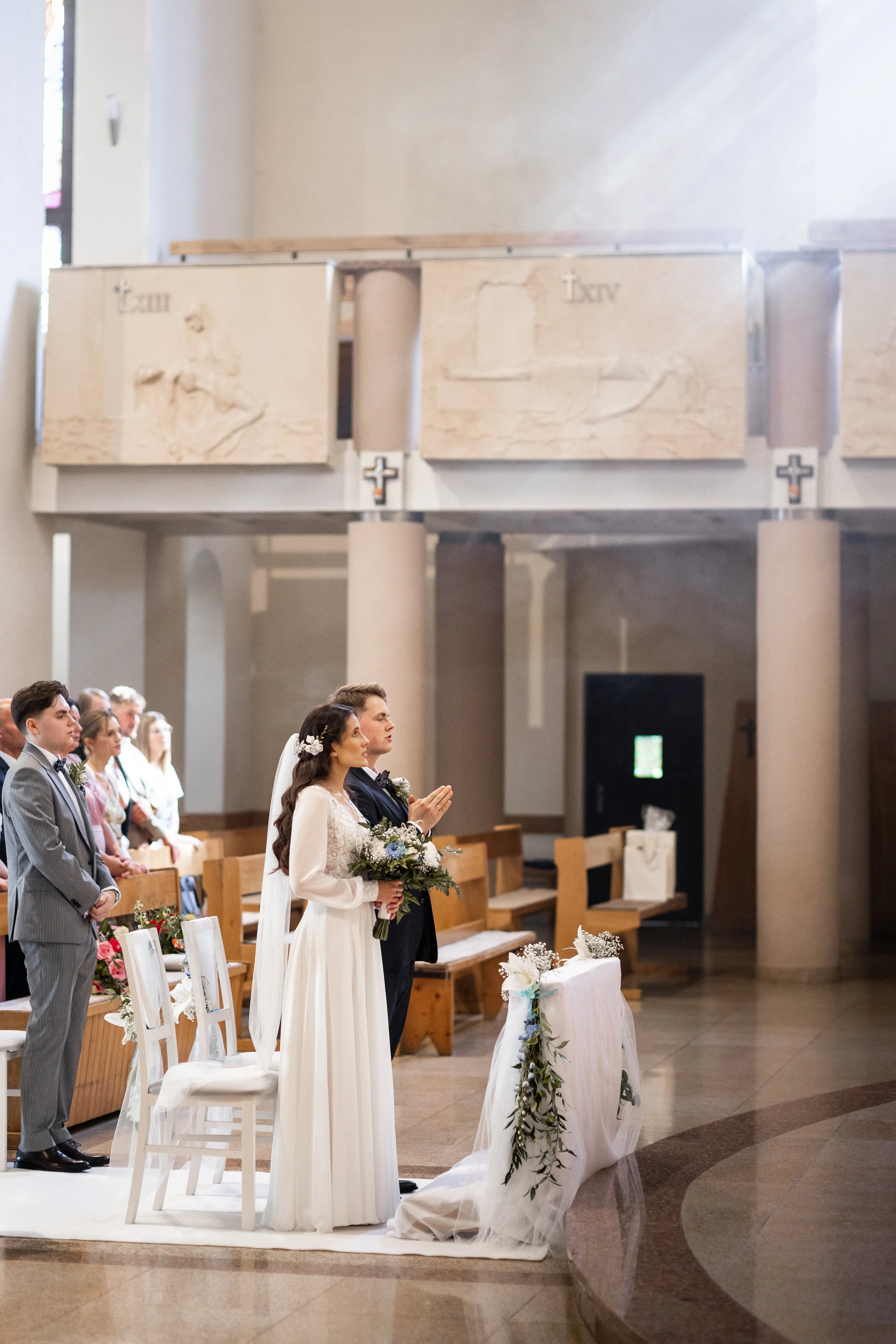 Para młoda stoi przy ołtarzu podczas ceremonii ślubnej w kościele, w tle siedzą goście weselni.