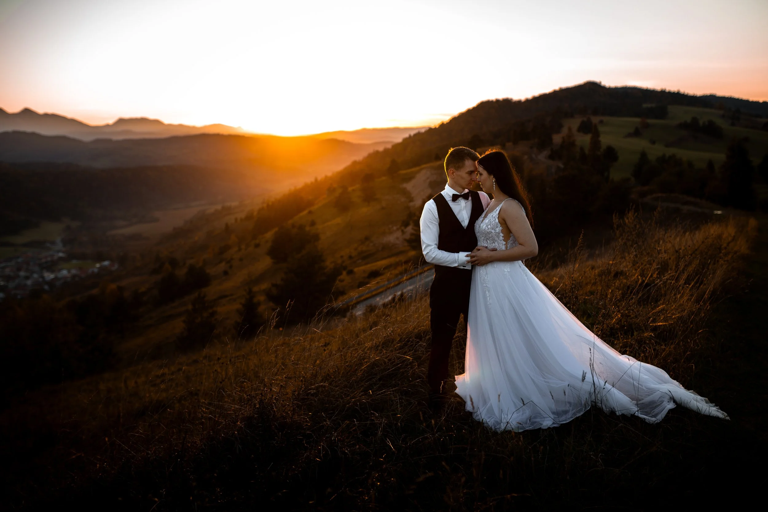 A bride and groom standing close together on a hillside during sunset, with mountains and a small village in the background.