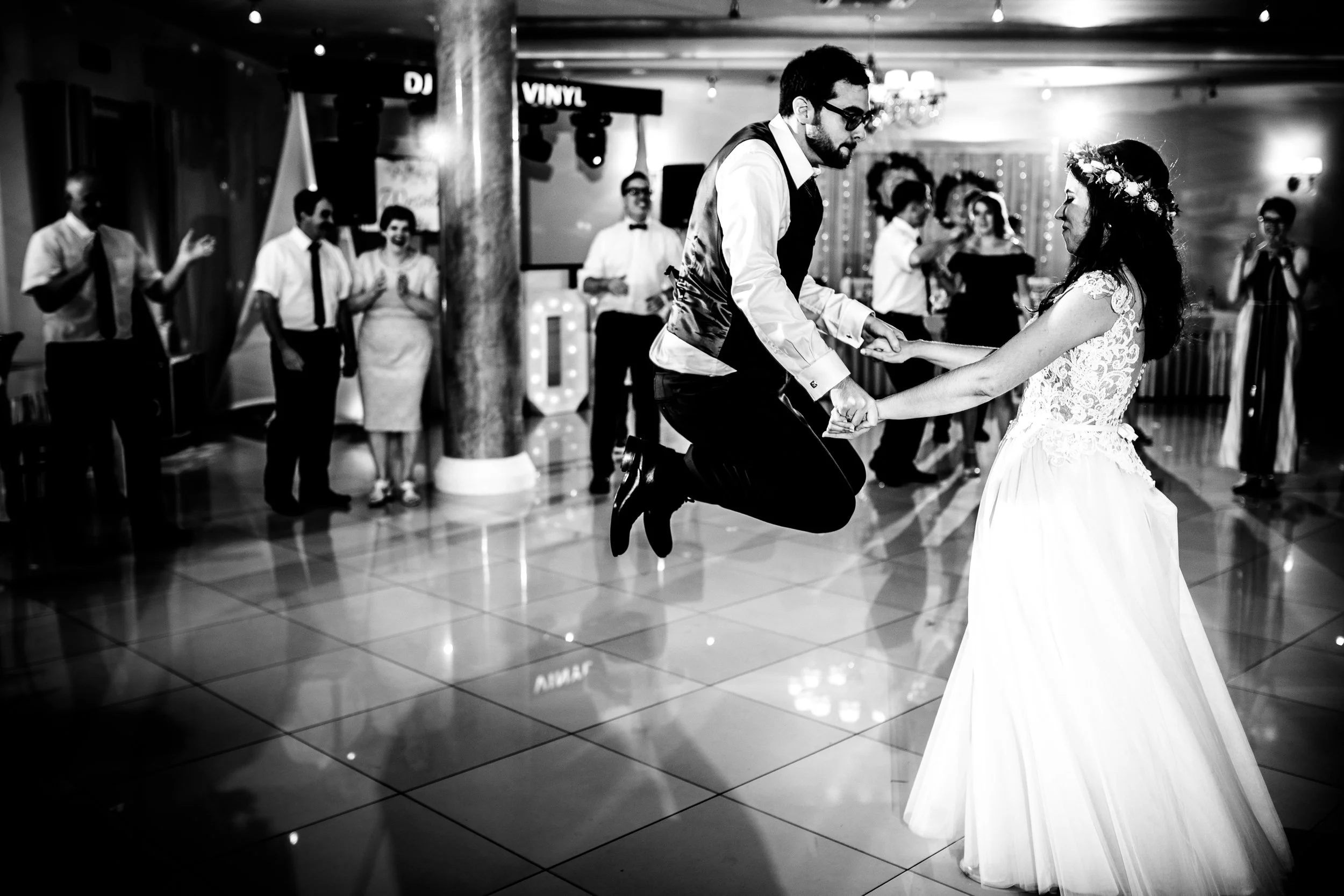 Black and white photo of a wedding reception dance floor with a groom and bride dancing together. The groom is jumping in the air while holding the bride's hands. The bride is wearing a long white gown with lace details and a floral crown. Several gu
