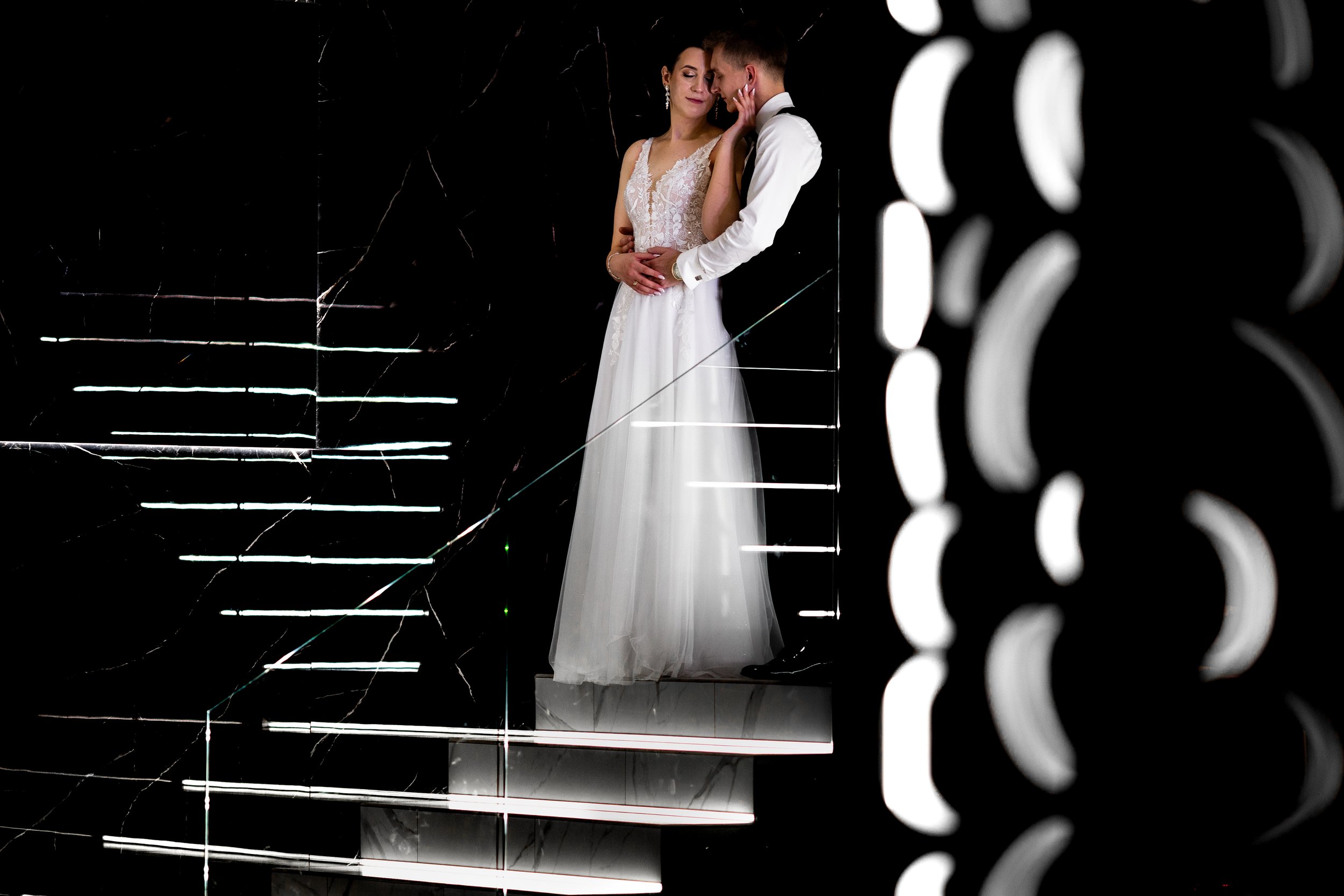 A bride and groom standing on a illuminated staircase, with the bride in a white wedding gown and the groom in a white shirt with dark pants, embracing each other in a dimly lit modern setting.