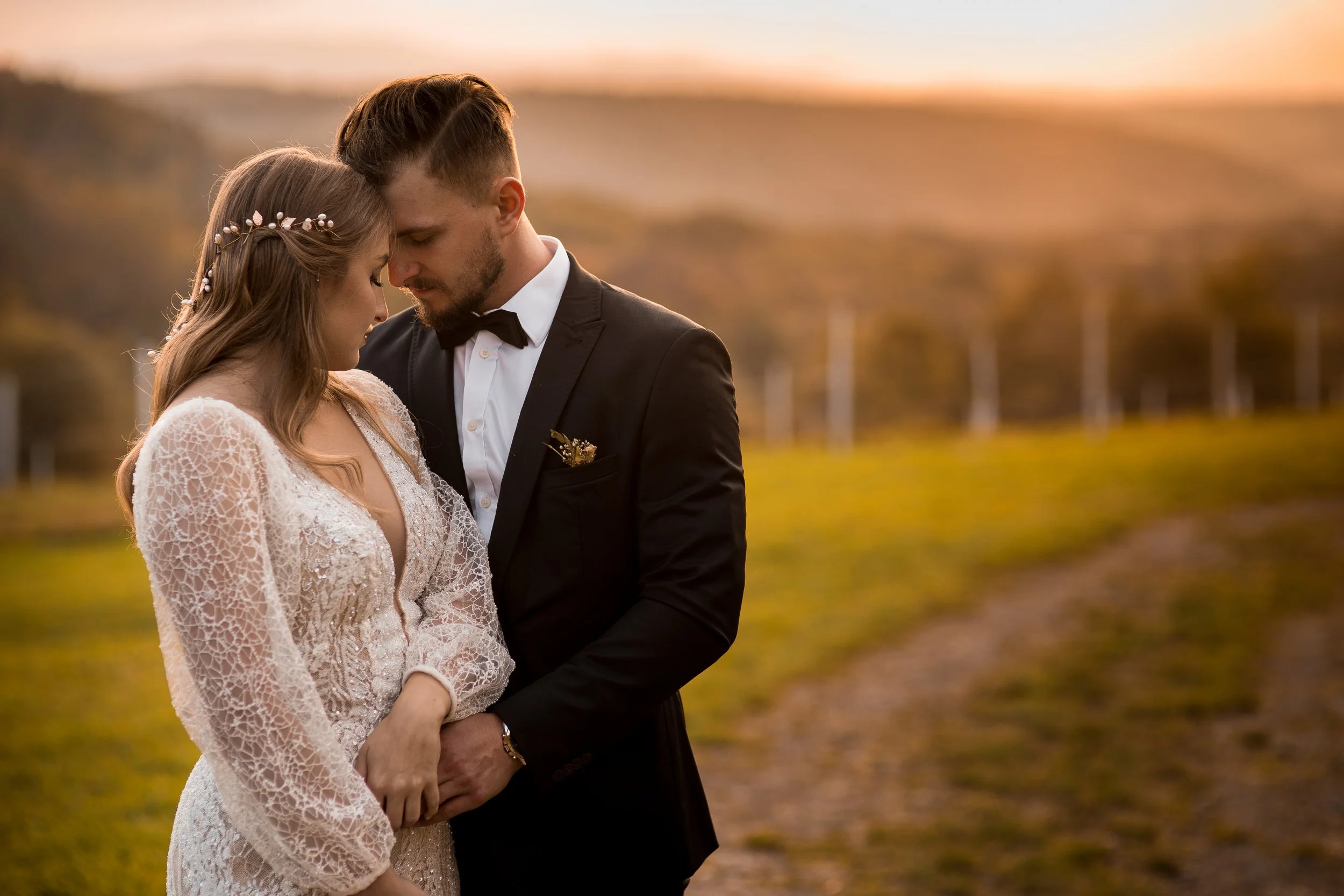 A bride and groom in an outdoor field at sunset, with their foreheads touching and eyes closed, dressed in wedding attire.