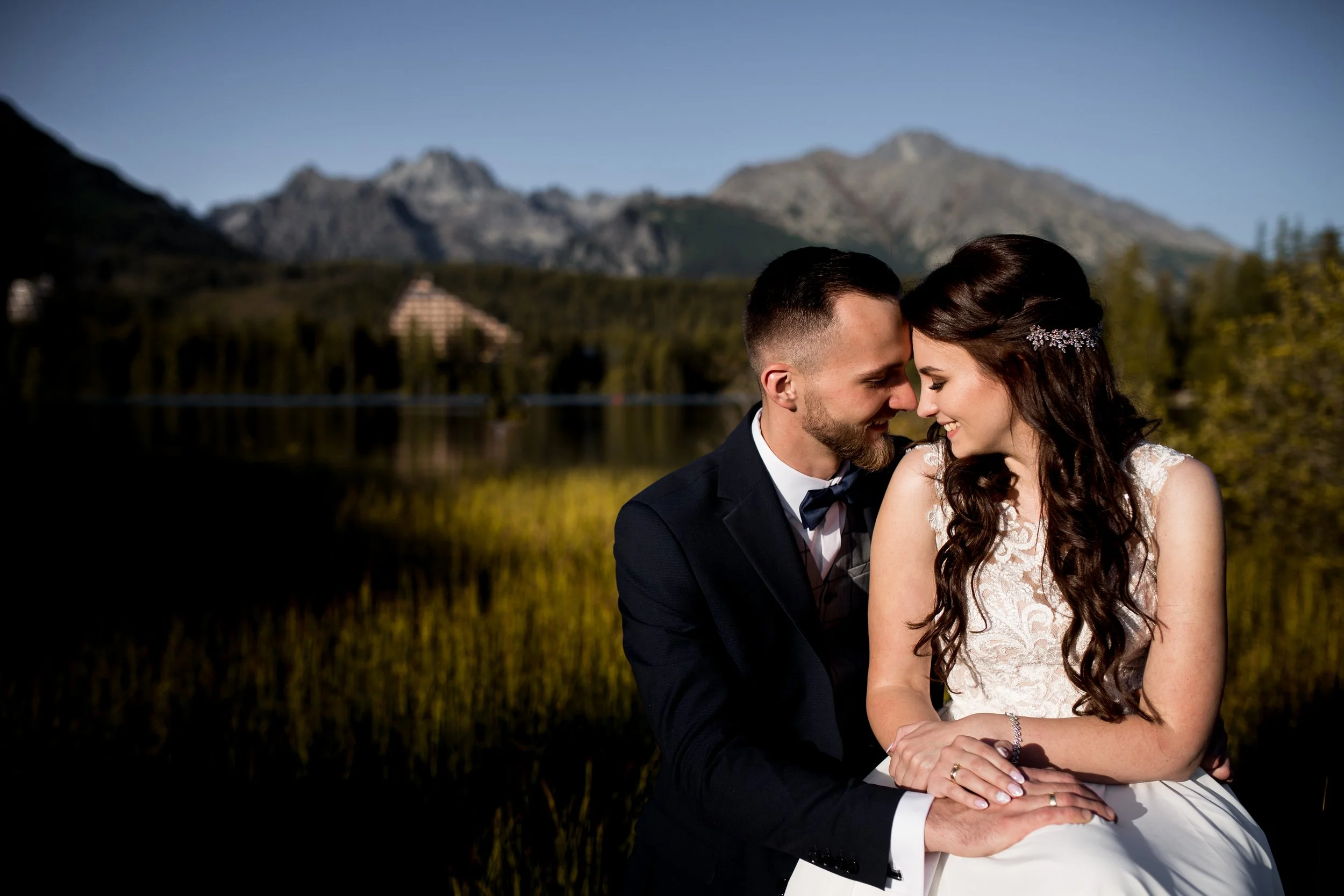 A bride and groom sitting close together outdoors near a lake with mountains in the background, smiling and touching foreheads.