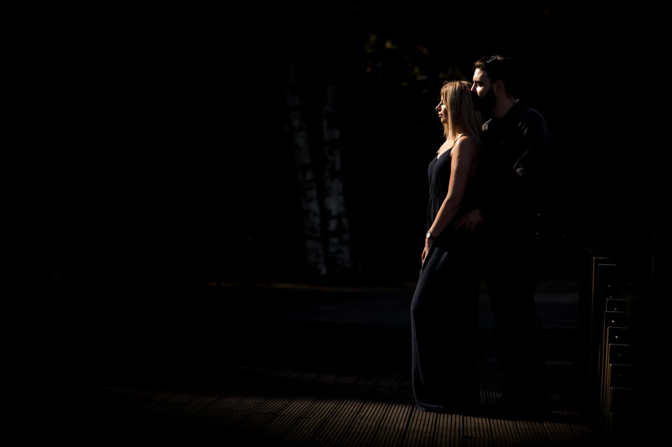 A couple stands close together outdoors at sunset, silhouetted against a dark background with a hint of trees in the distance.