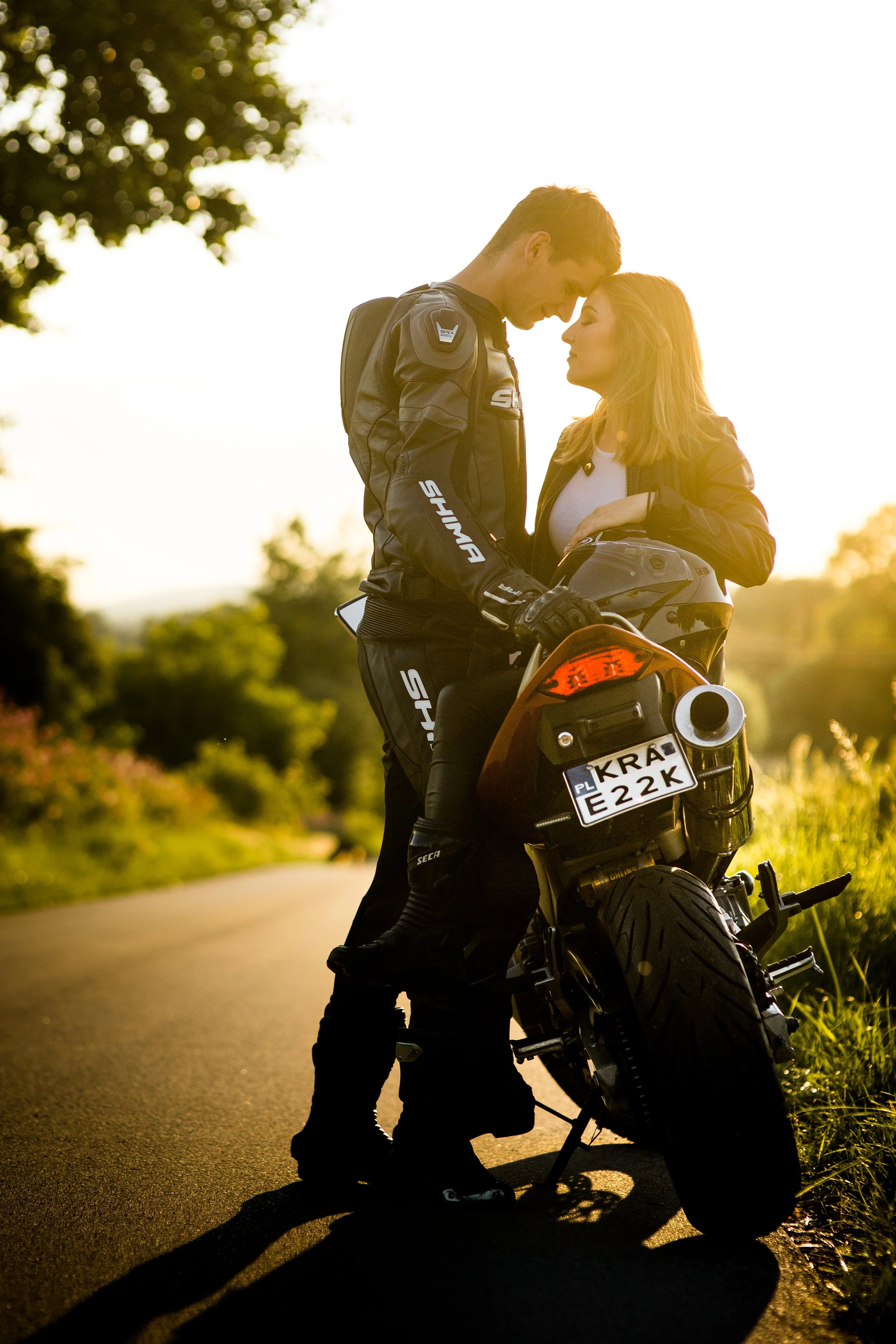 Młody mężczyzna i kobieta stoją blisko siebie obok motocykla na wiejskiej drodze o zachodzie słońca. Mężczyzna ubrany jest w strój motocyklowy, a kobieta w codziennym ubraniu, stykając się czołami.
