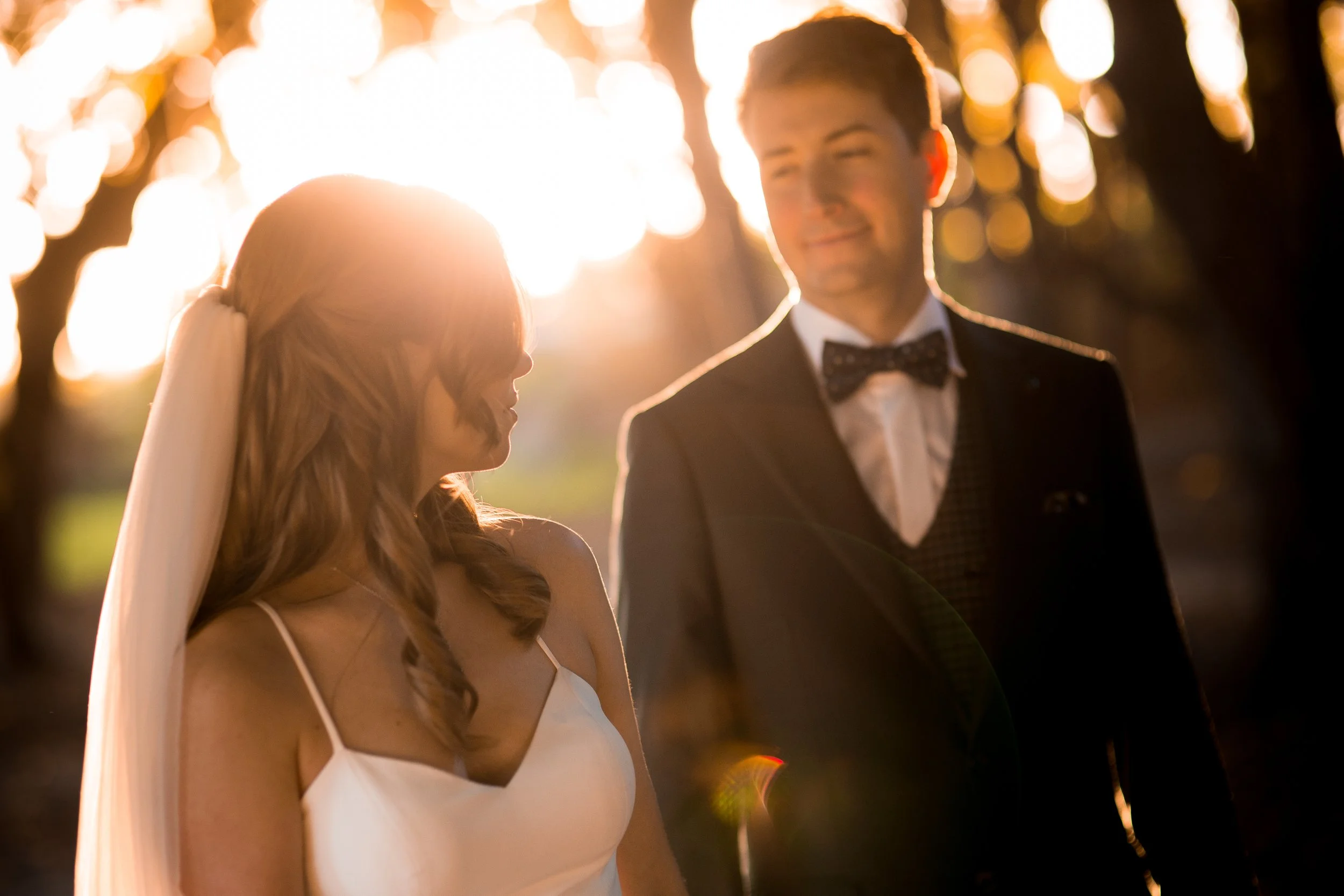 A couple dressed in wedding attire stands outdoors during sunset. The bride has long, wavy red hair, and the groom has dark hair and is wearing a tuxedo with a bow tie. The sunlight creates a warm, glowing backlight around them.