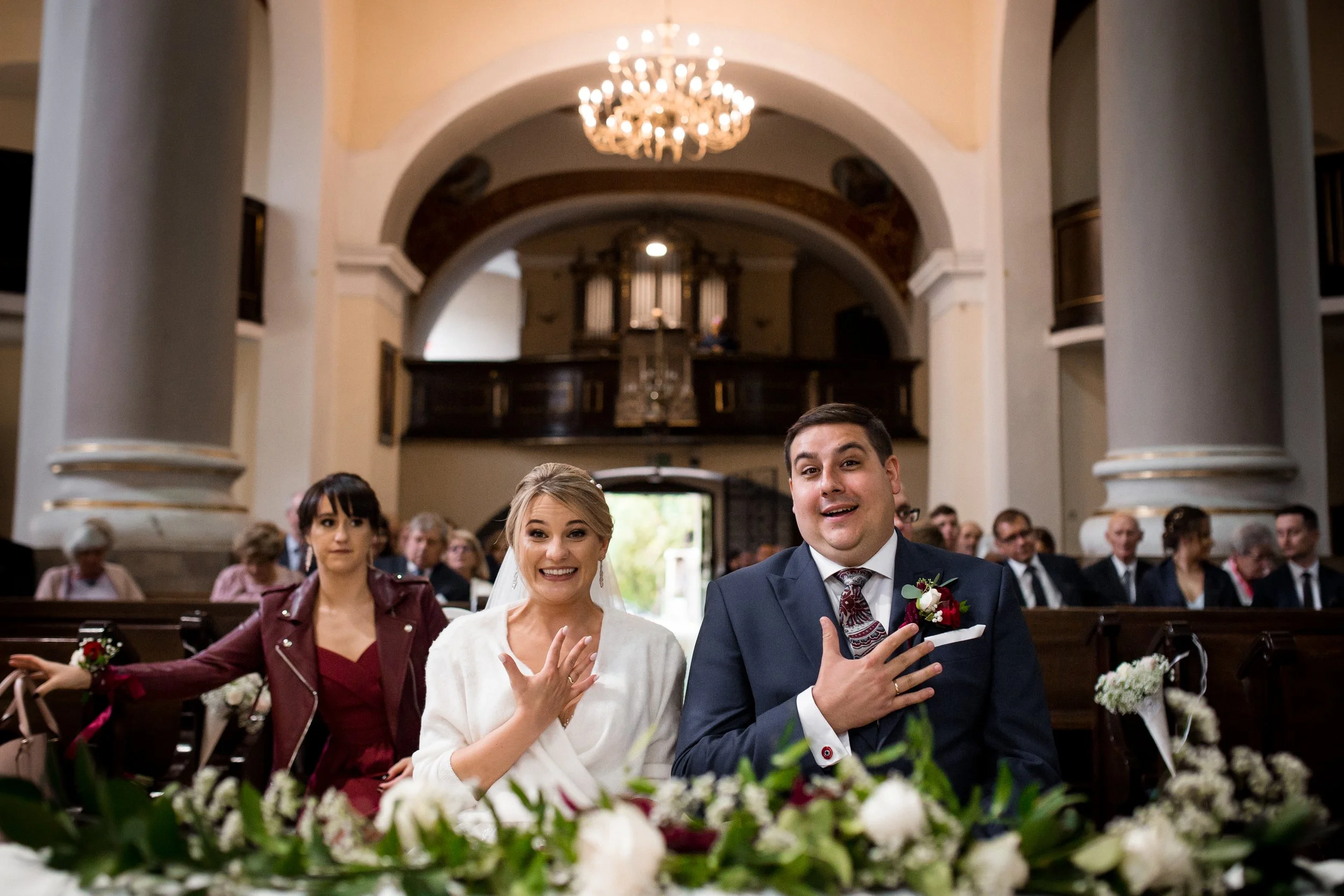 Wedding ceremony inside a church with three people in the foreground performing a ritual, surrounded by guests, floral arrangements, and a large chandelier overhead.