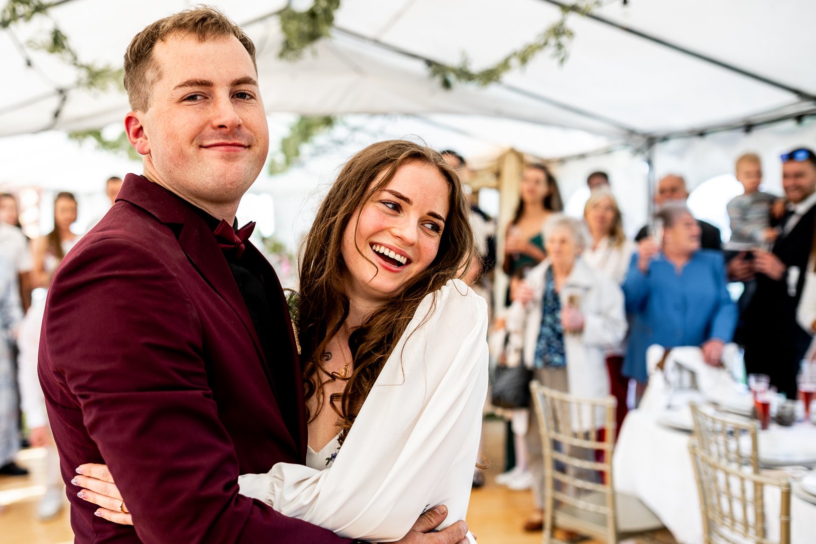 A couple at a wedding reception with the groom in a burgundy tuxedo and the bride in a white dress, smiling and embracing.