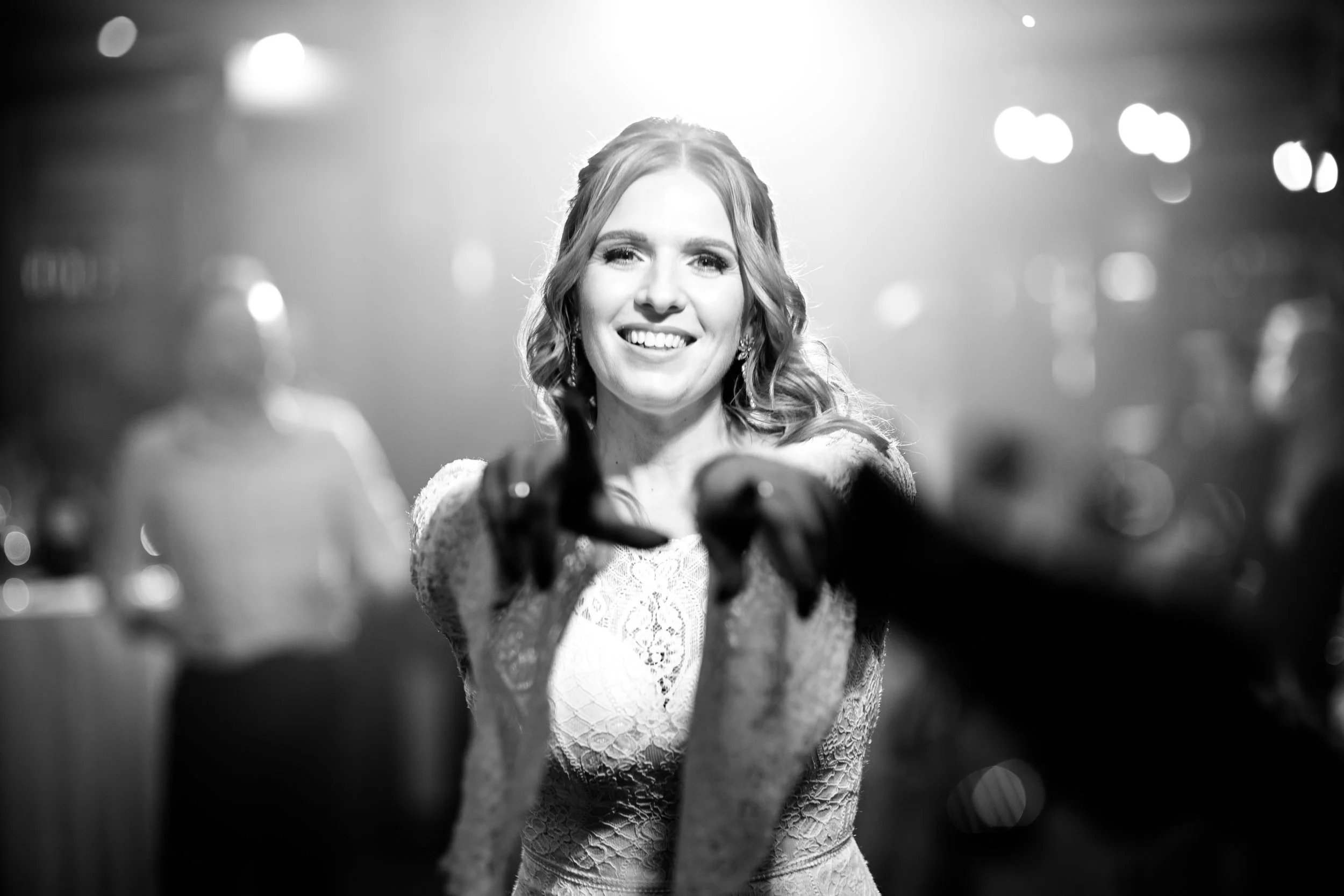 A smiling woman with wavy hair in a lace dress reaching out towards the camera, with a blurred background and bright lights.