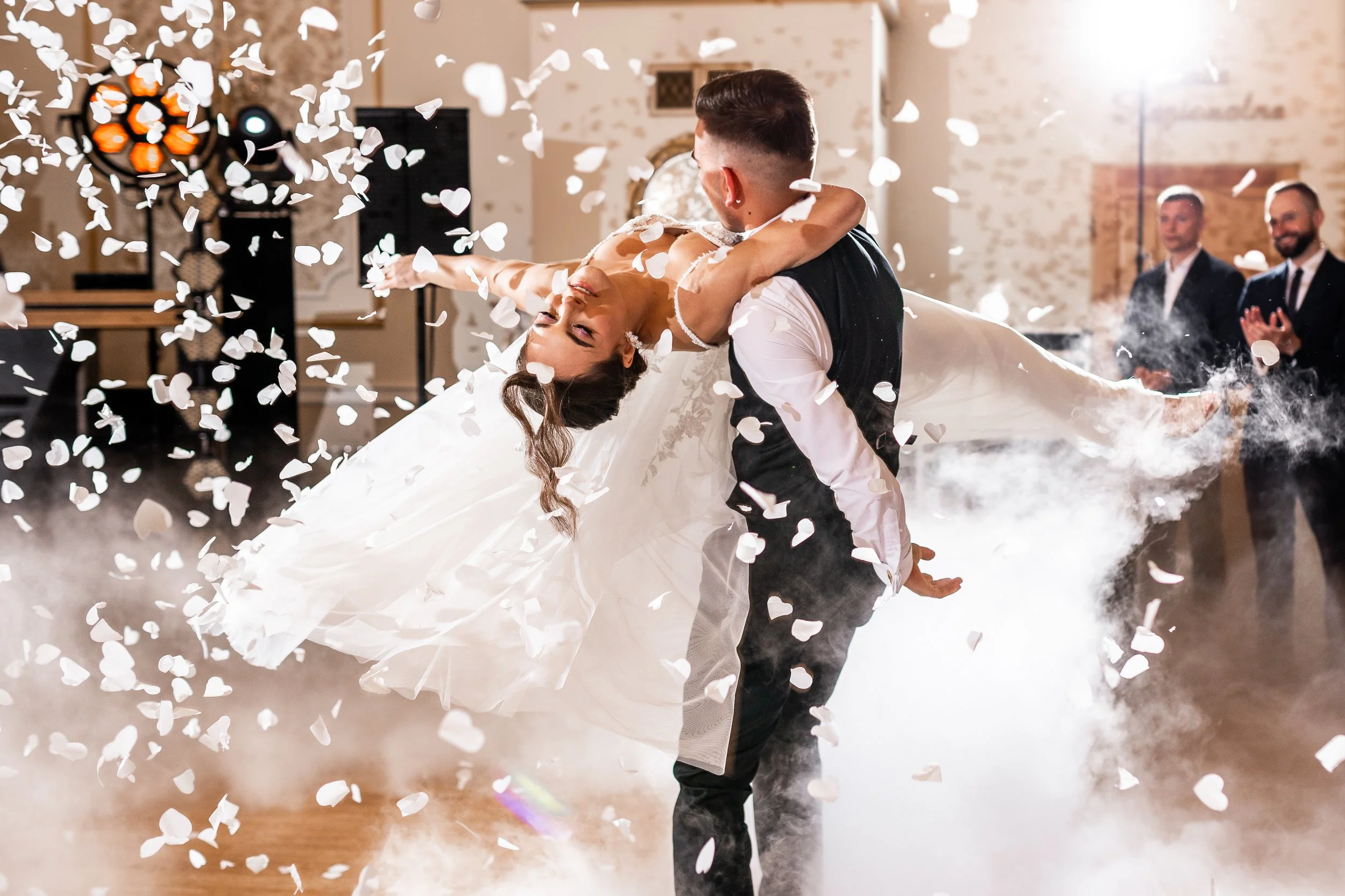 A newlywed couple shares their first dance at a wedding reception, with the groom dipping the bride while floating white heart-shaped confetti around them. The background features wedding guests clapping and cheering.