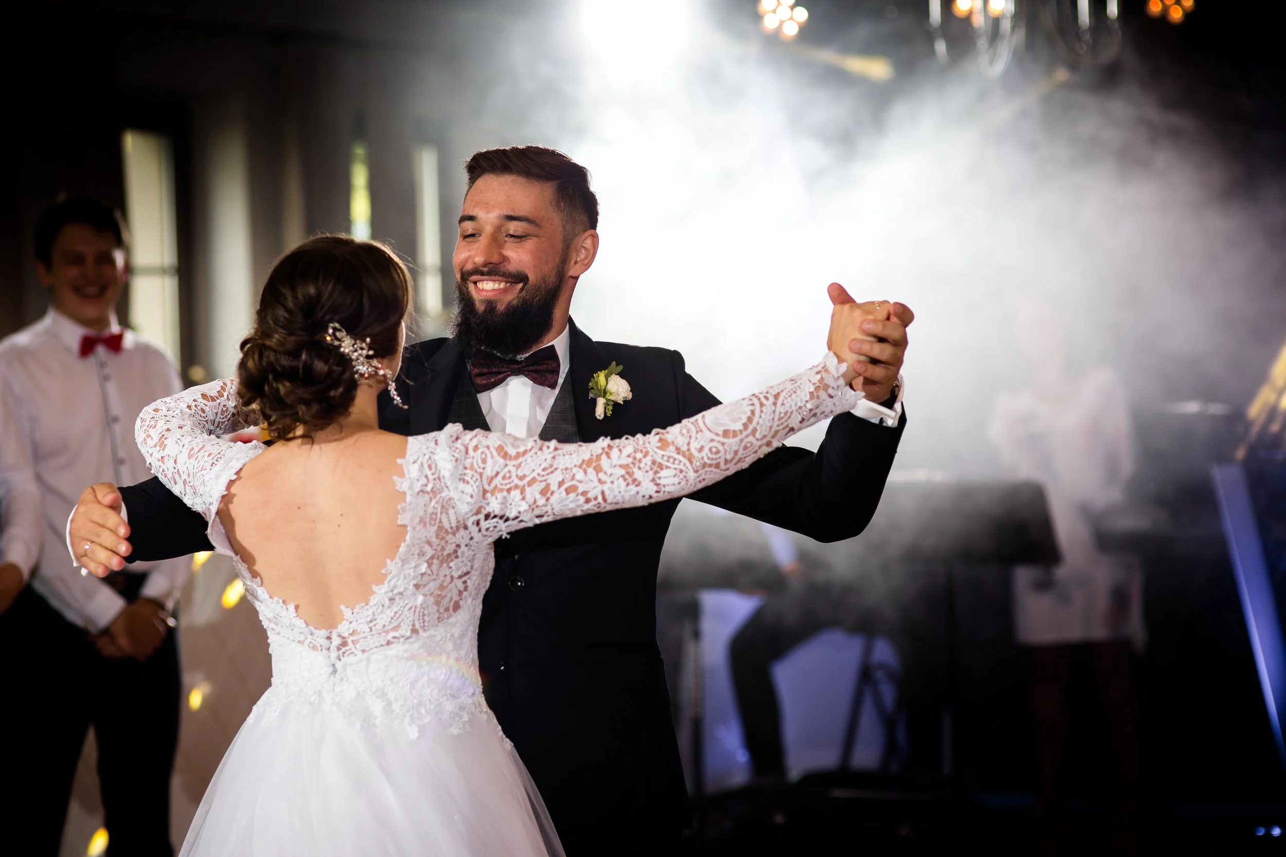 A bride and groom dancing at their wedding reception, with a guest in the background smiling.