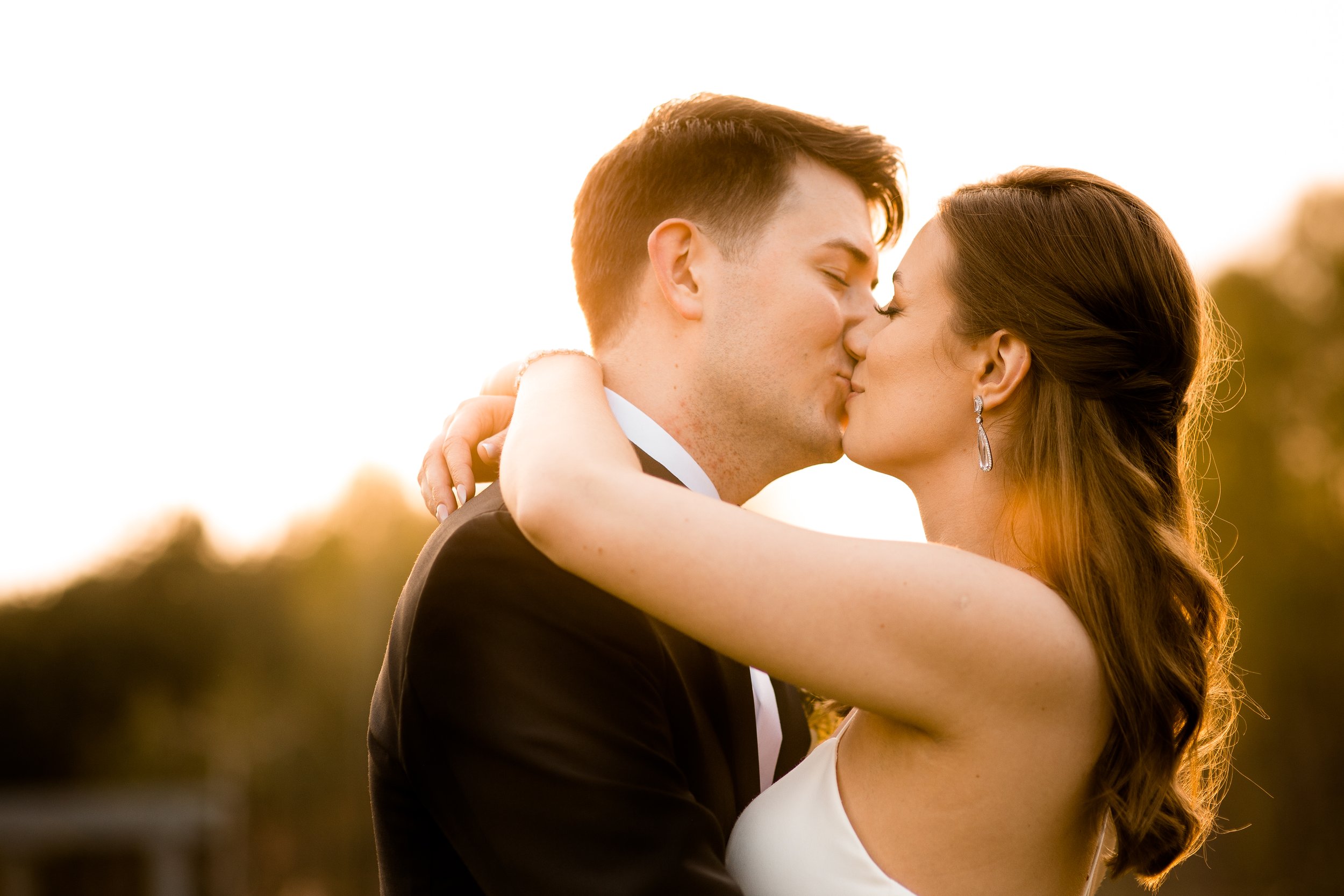 A bride and groom kissing outdoors during sunset, embracing each other with eyes closed.