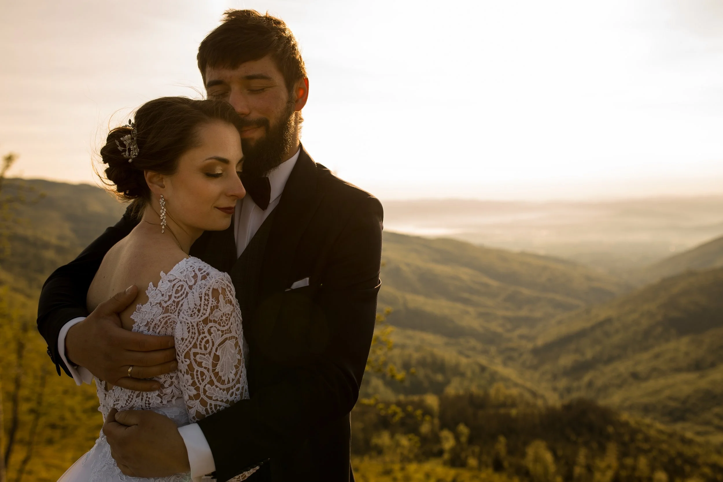 A newlywed couple hugging outdoors during sunset, with rolling hills in the background.