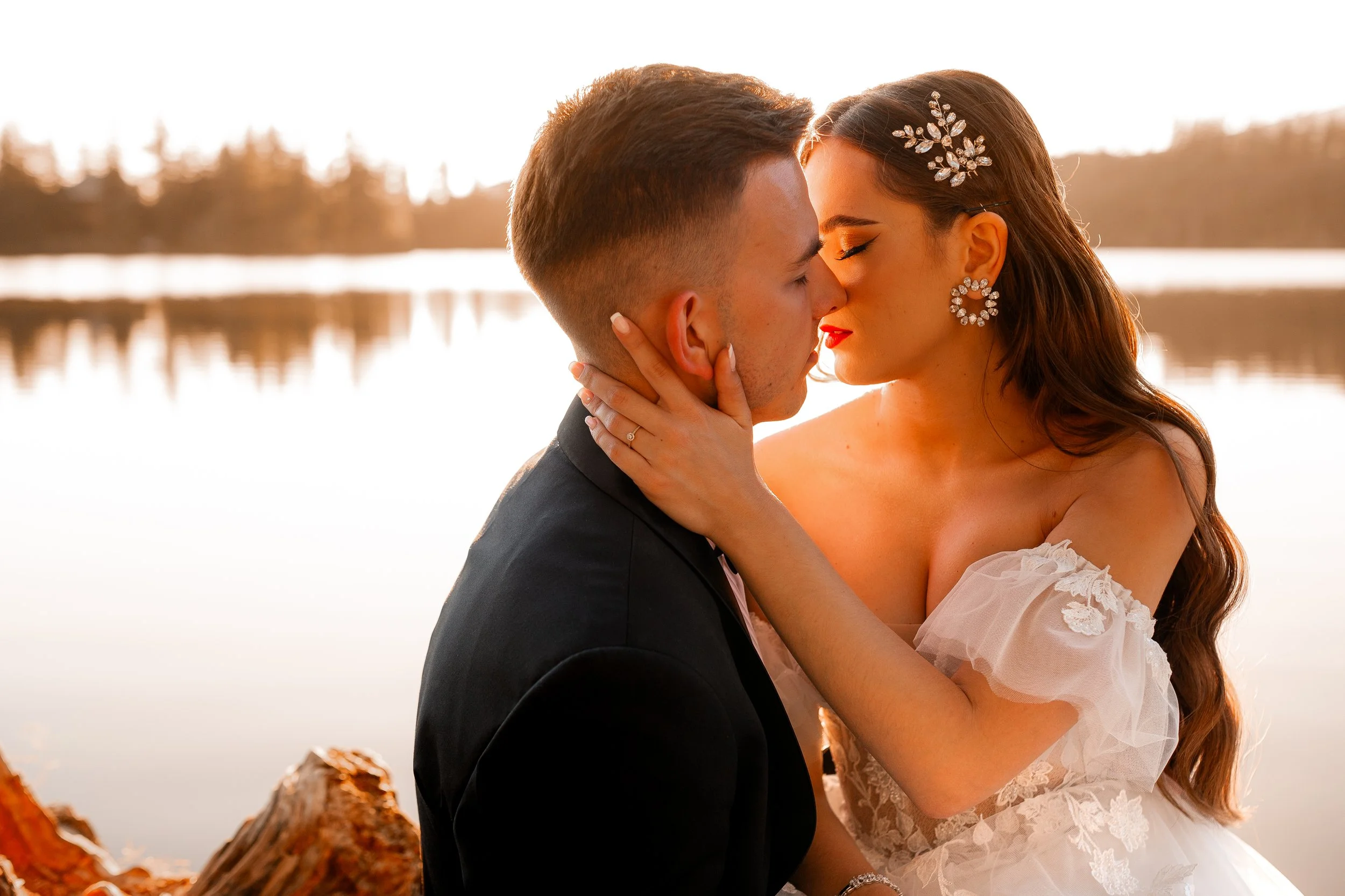 A bride and groom sharing a kiss near a lake at sunset, with trees in the background.