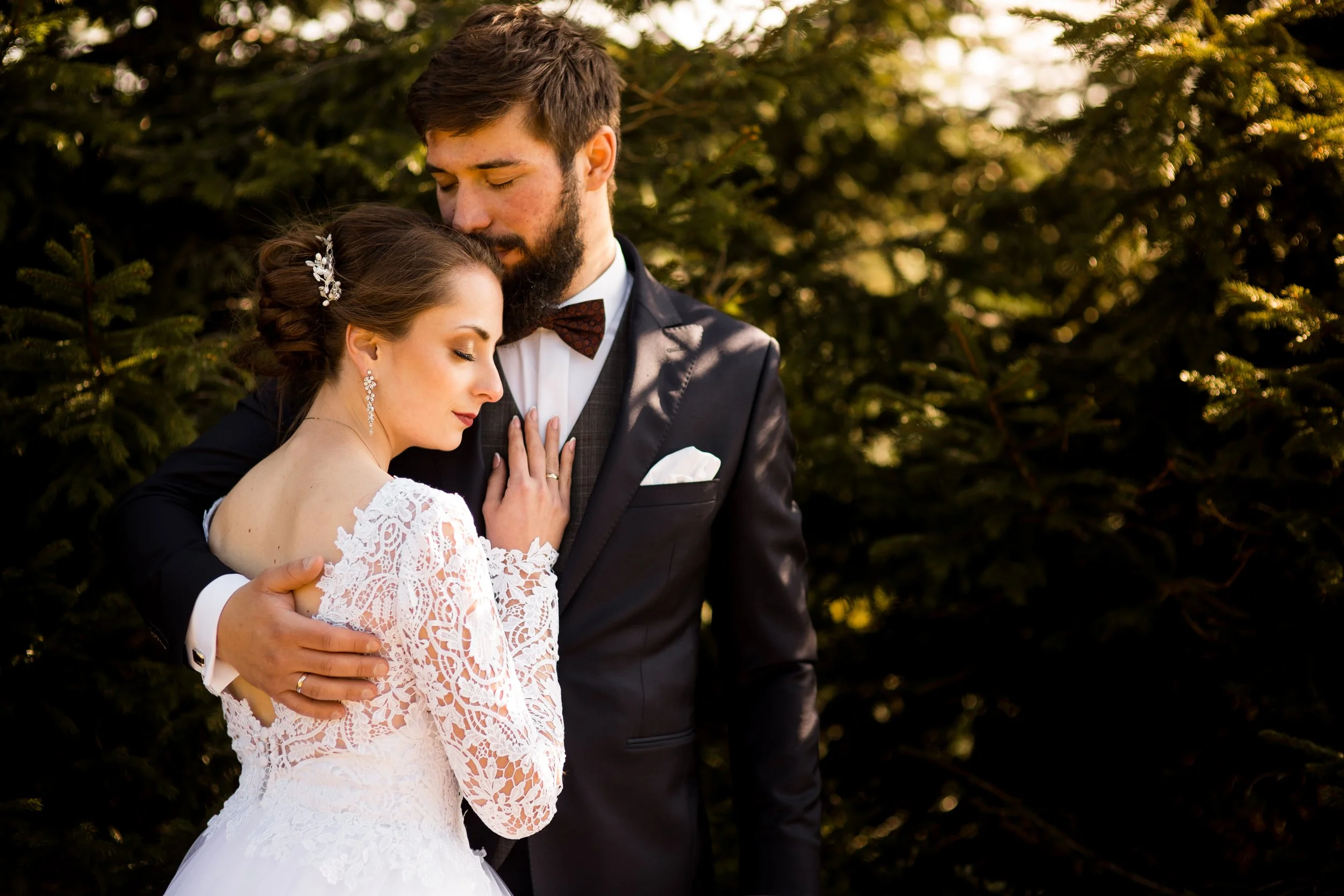 A newlywed couple embraces outdoors in front of trees, with the bride in a white lace wedding dress and the groom in a dark suit with a bow tie.
