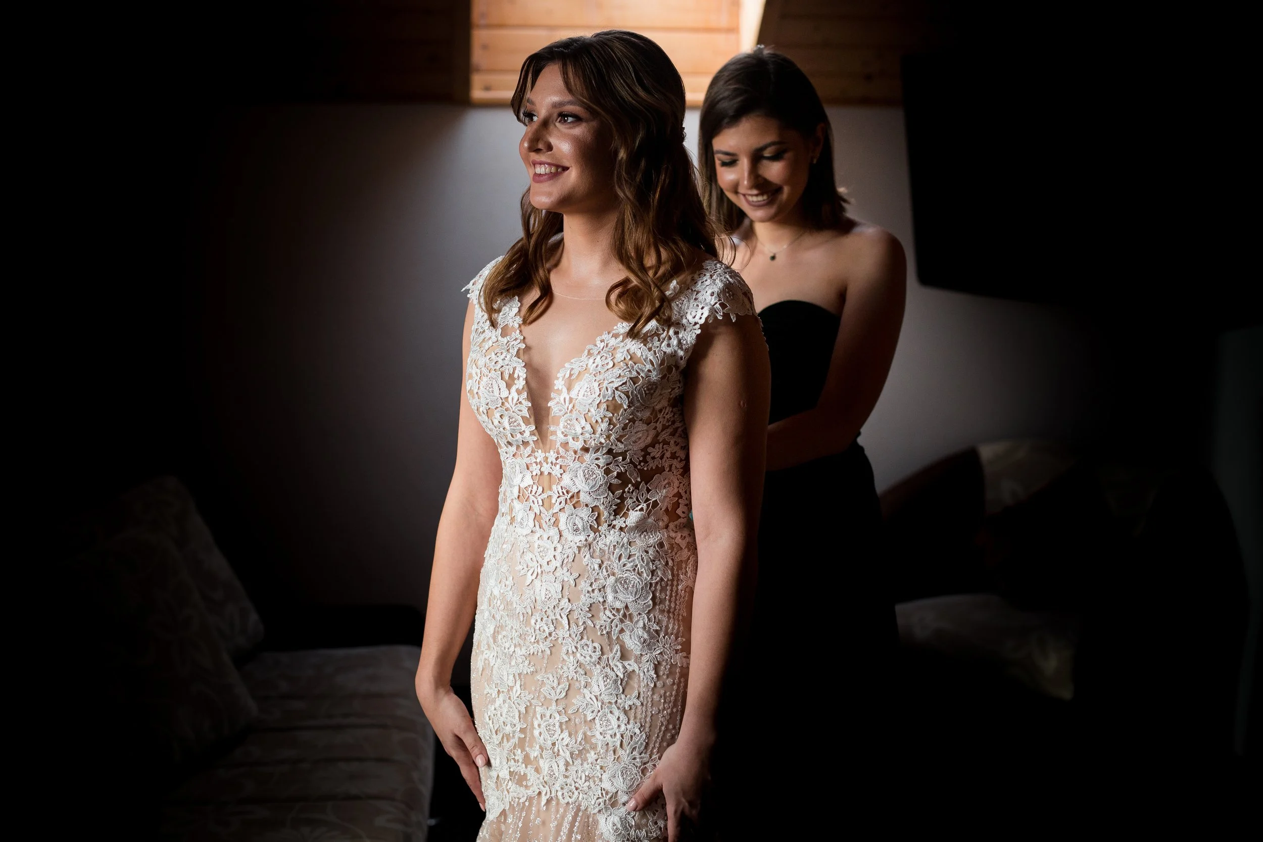 A bride in a lace wedding dress being assisted by a bridesmaid in a black dress.