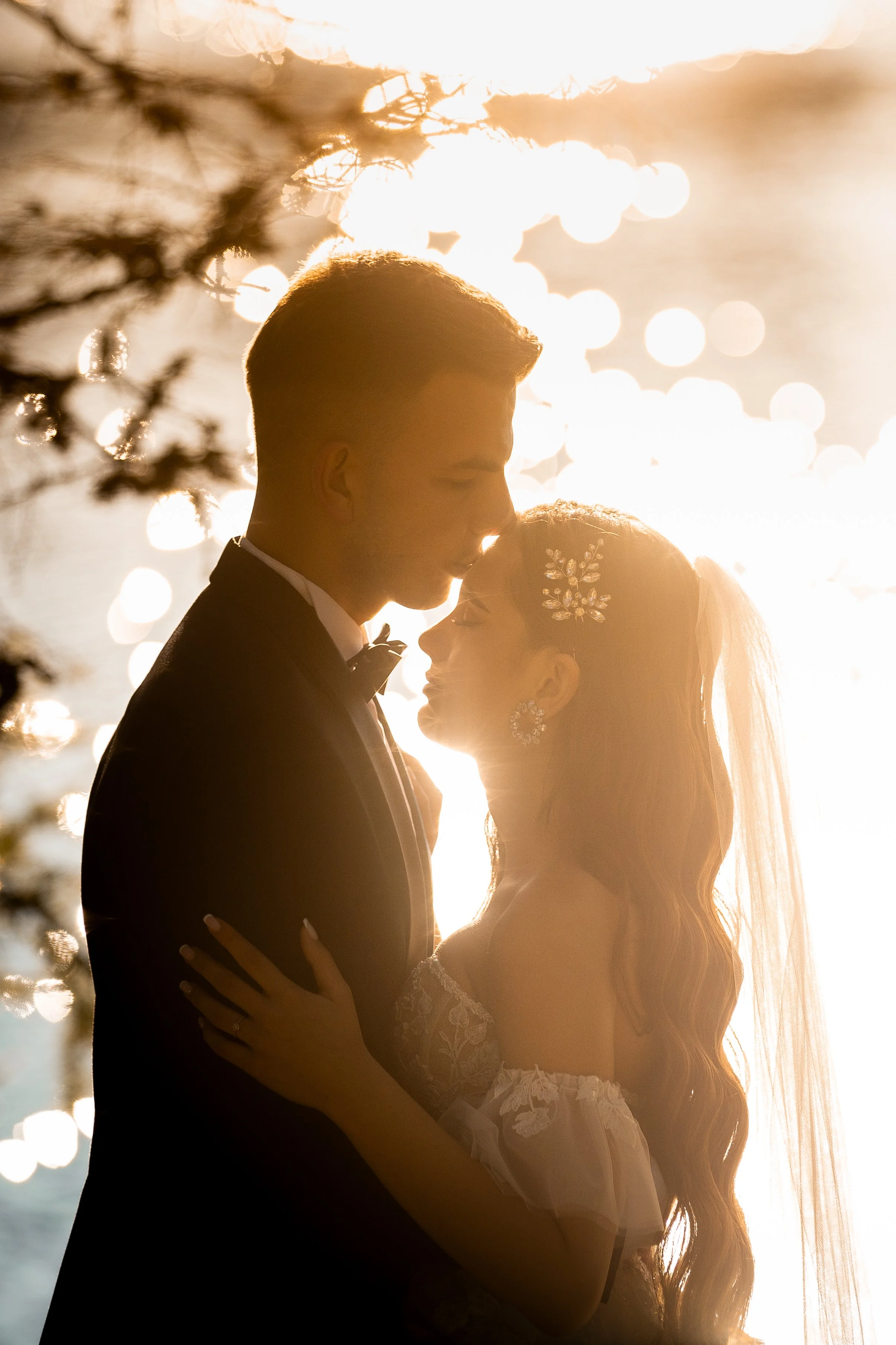 Silhouette of a bride and groom embracing, backlit by warm sunlight reflecting on the water, with trees in the foreground.