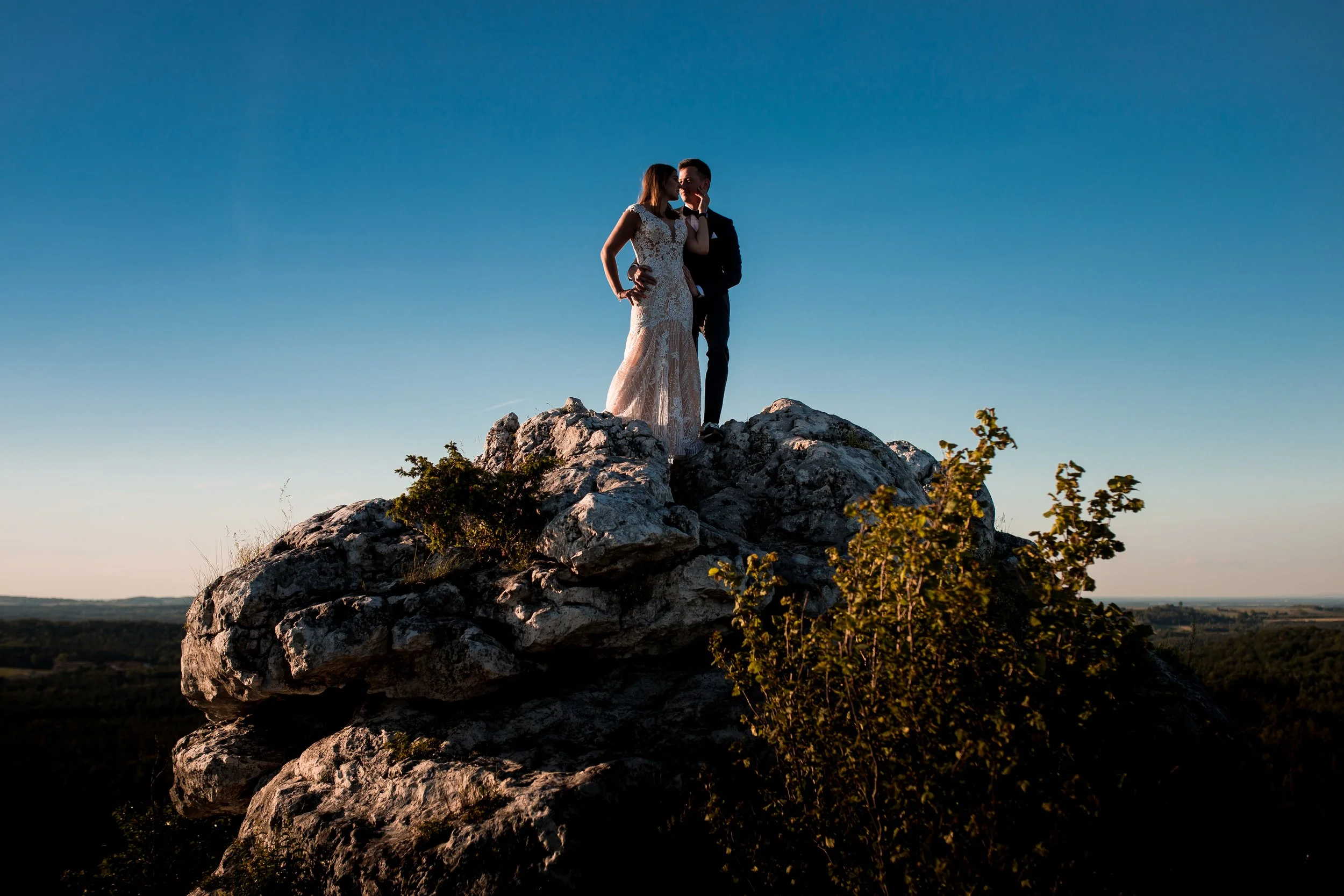 A couple in wedding attire standing on a rocky outcrop during sunset, with a clear blue sky in the background.