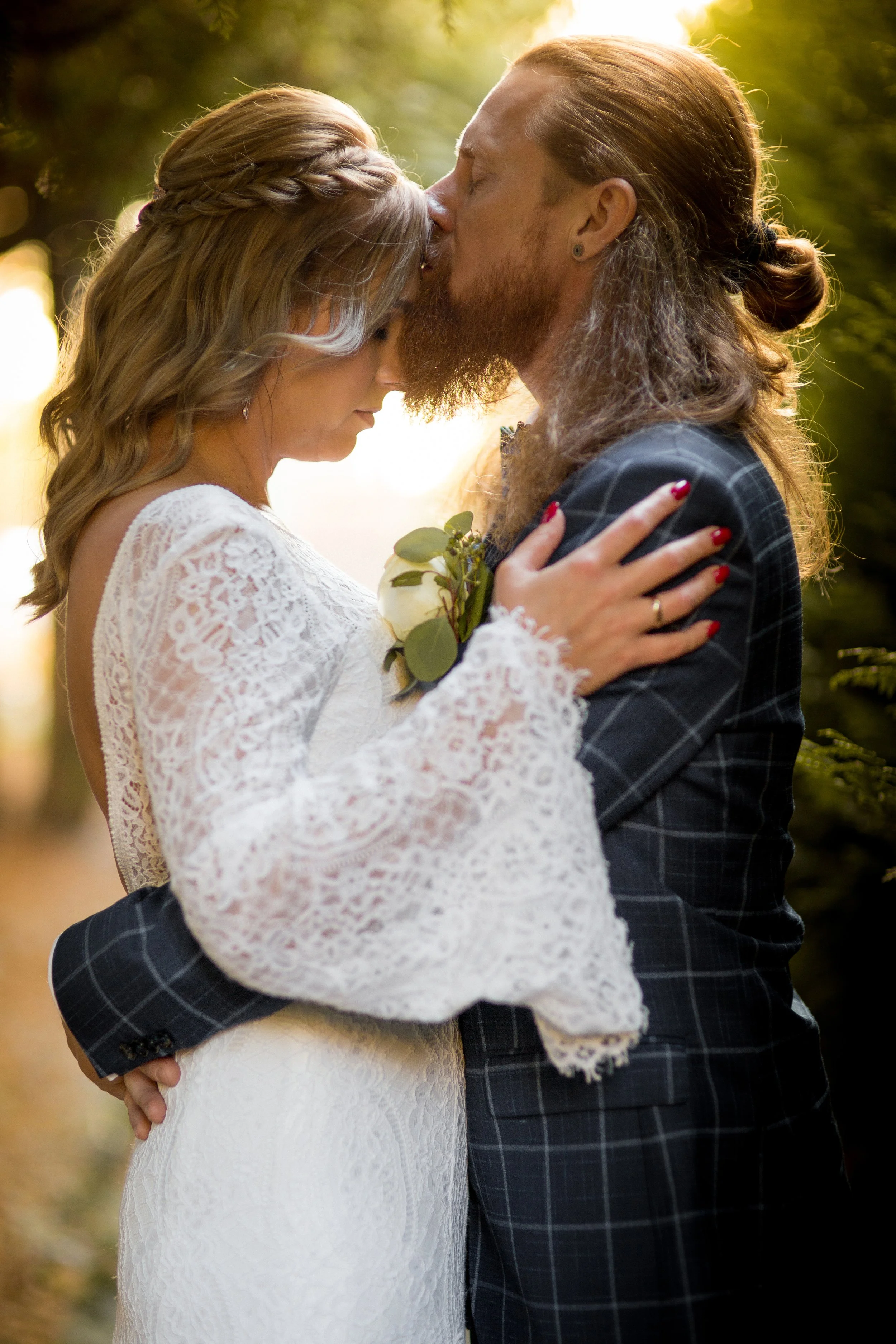 A couple in wedding attire sharing an intimate moment outdoors, with the man kissing the woman's forehead, both with eyes closed. The woman is wearing a white lace dress with long sleeves and a braided hairstyle, and the man has long hair, a beard, a