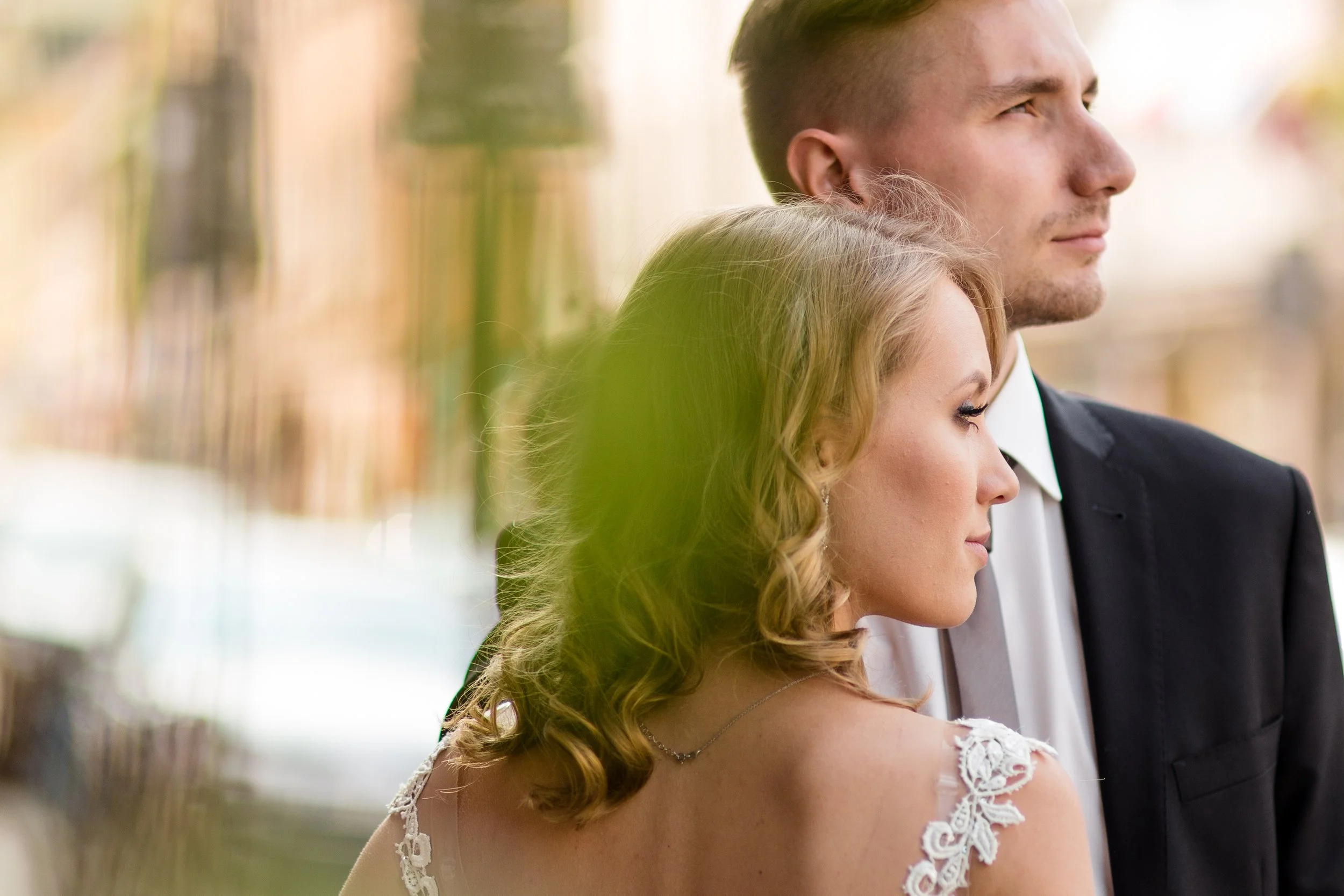 A bride and groom standing close together outdoors, facing right, with the bride slightly in front. The bride has curly blonde hair and is wearing a white lace wedding dress. The groom has short brown hair, light beard, and is wearing a black suit wi
