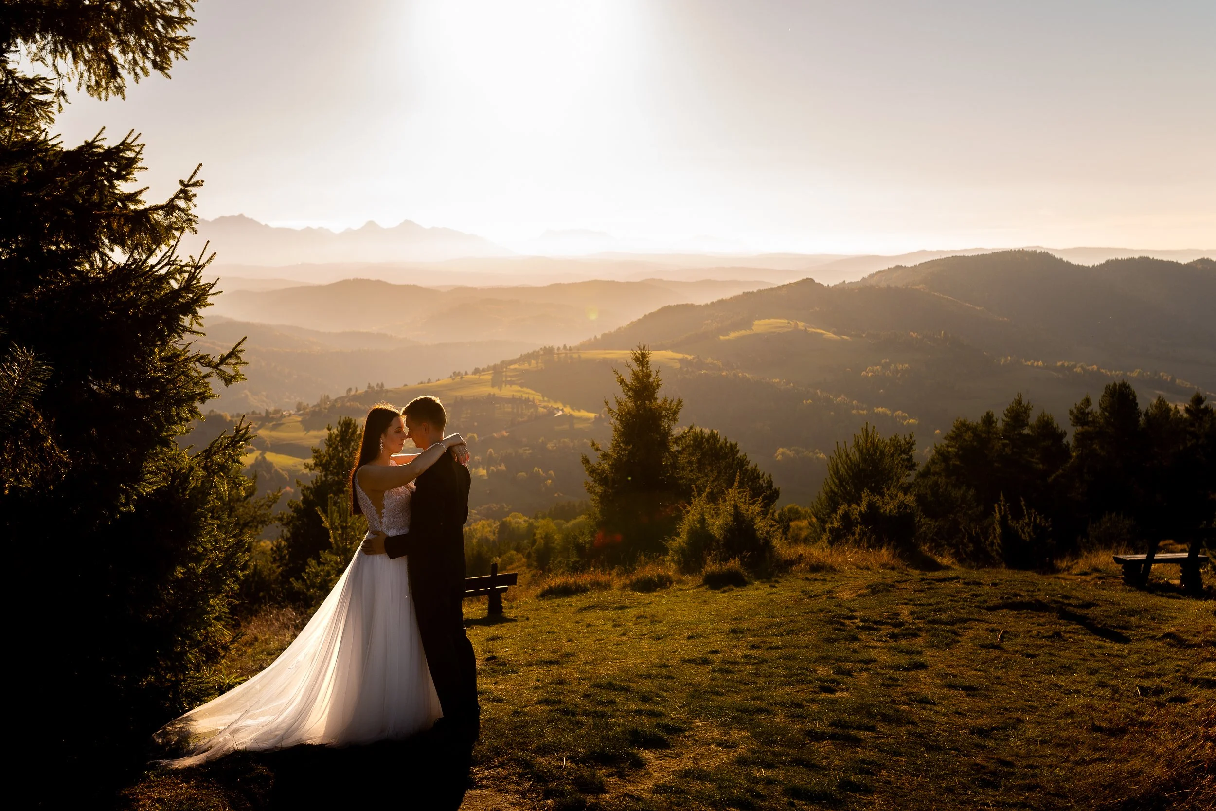 A bride and groom embracing outdoors at sunset, with a scenic mountain landscape in the background.