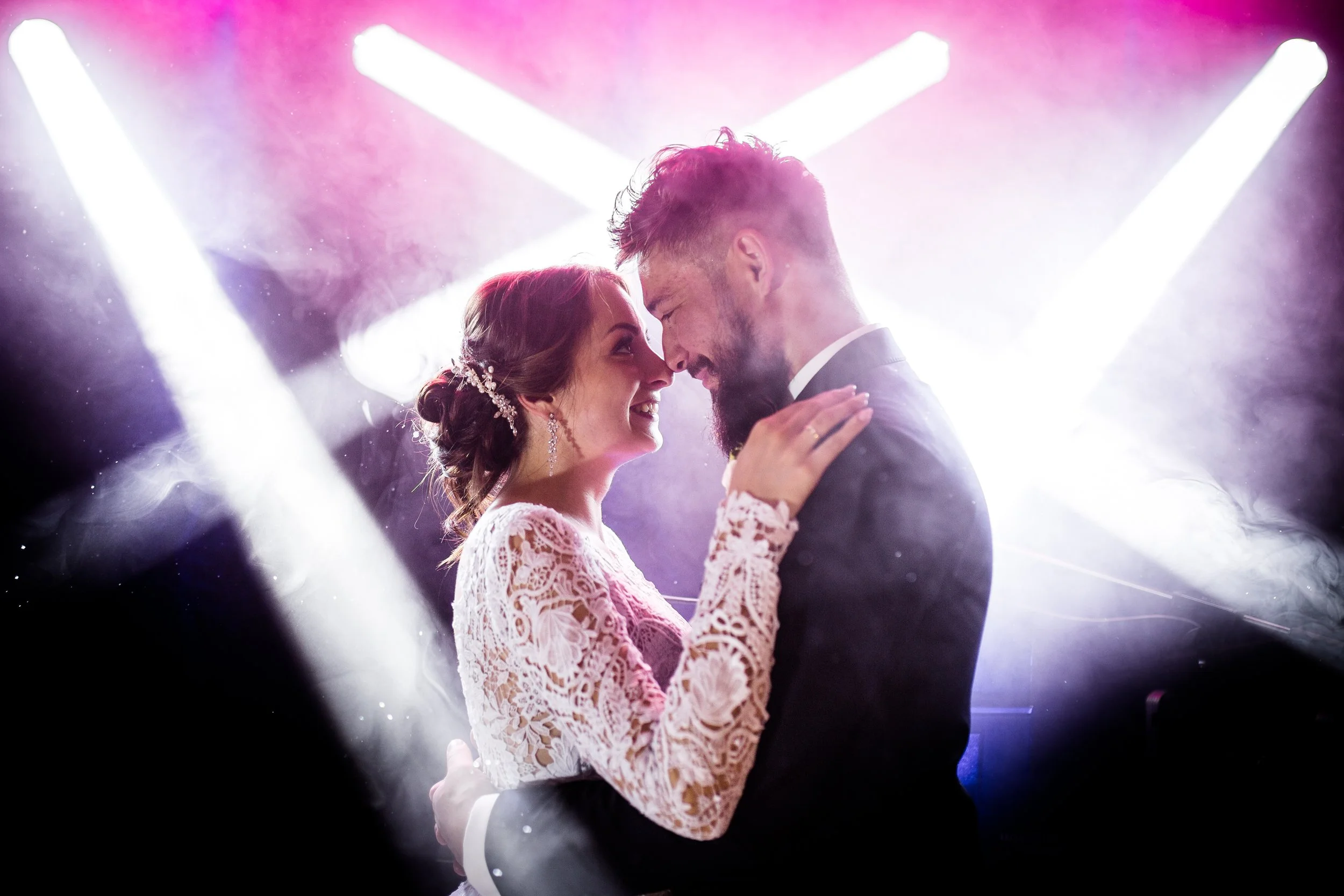 A bride and groom dancing close together at their wedding, smiling and touching foreheads, with bright lights and foggy atmosphere in the background.