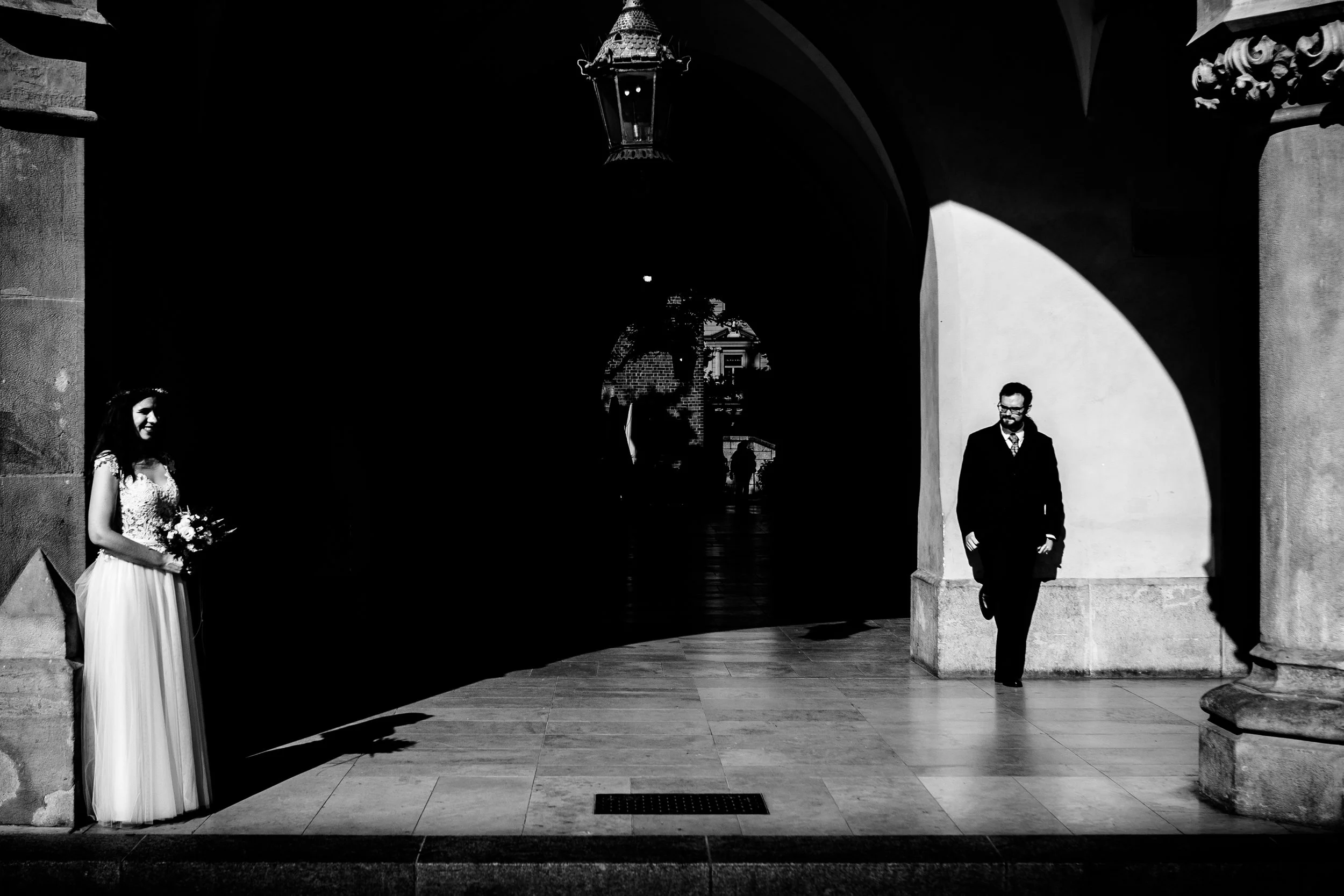 Black and white photo of a bride holding a bouquet on the left and a groom in a suit on the right, standing under an archway with shadows and bright sunlight.