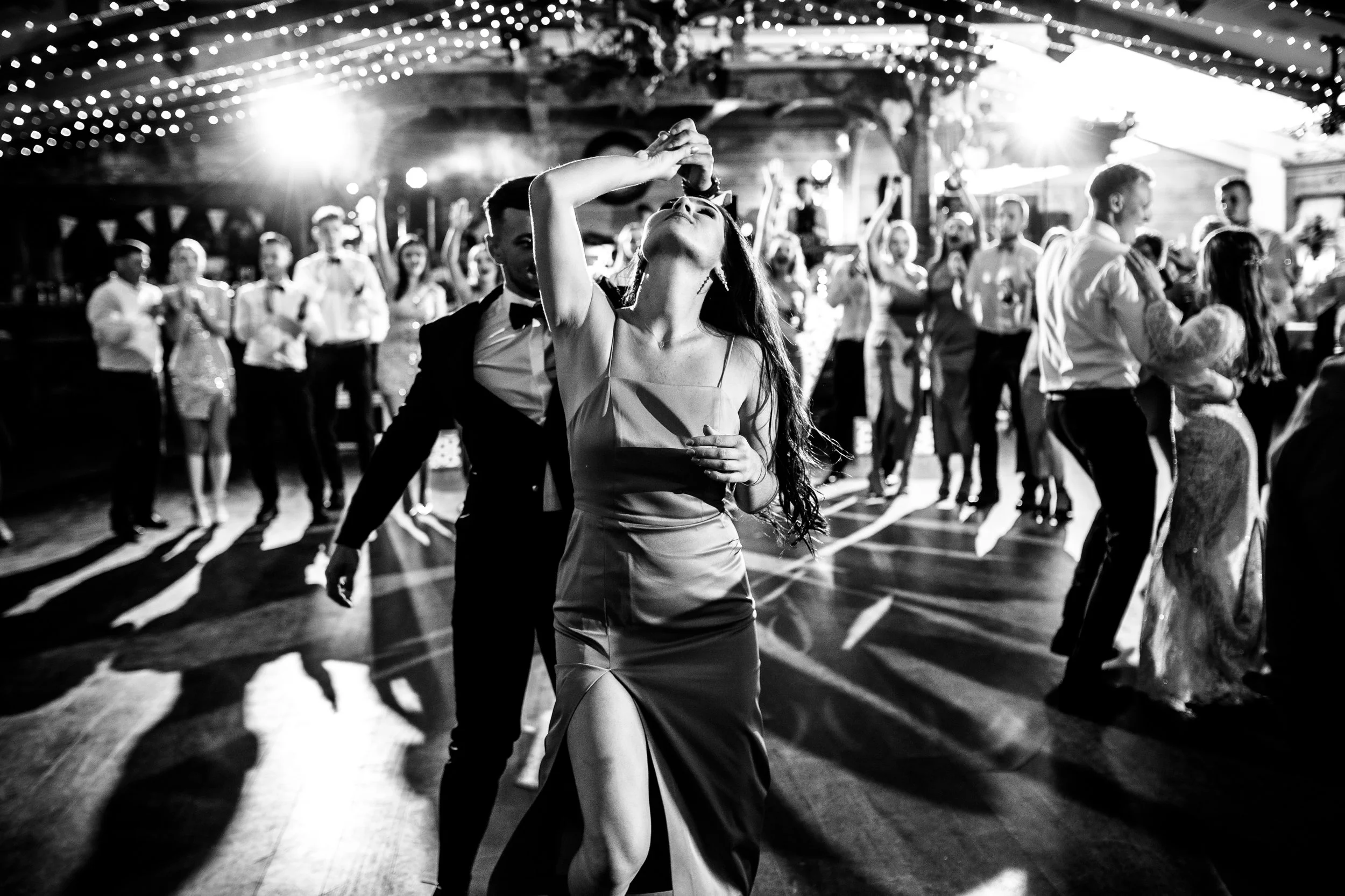 People dancing at a wedding reception in a decorated venue with string lights, black and white