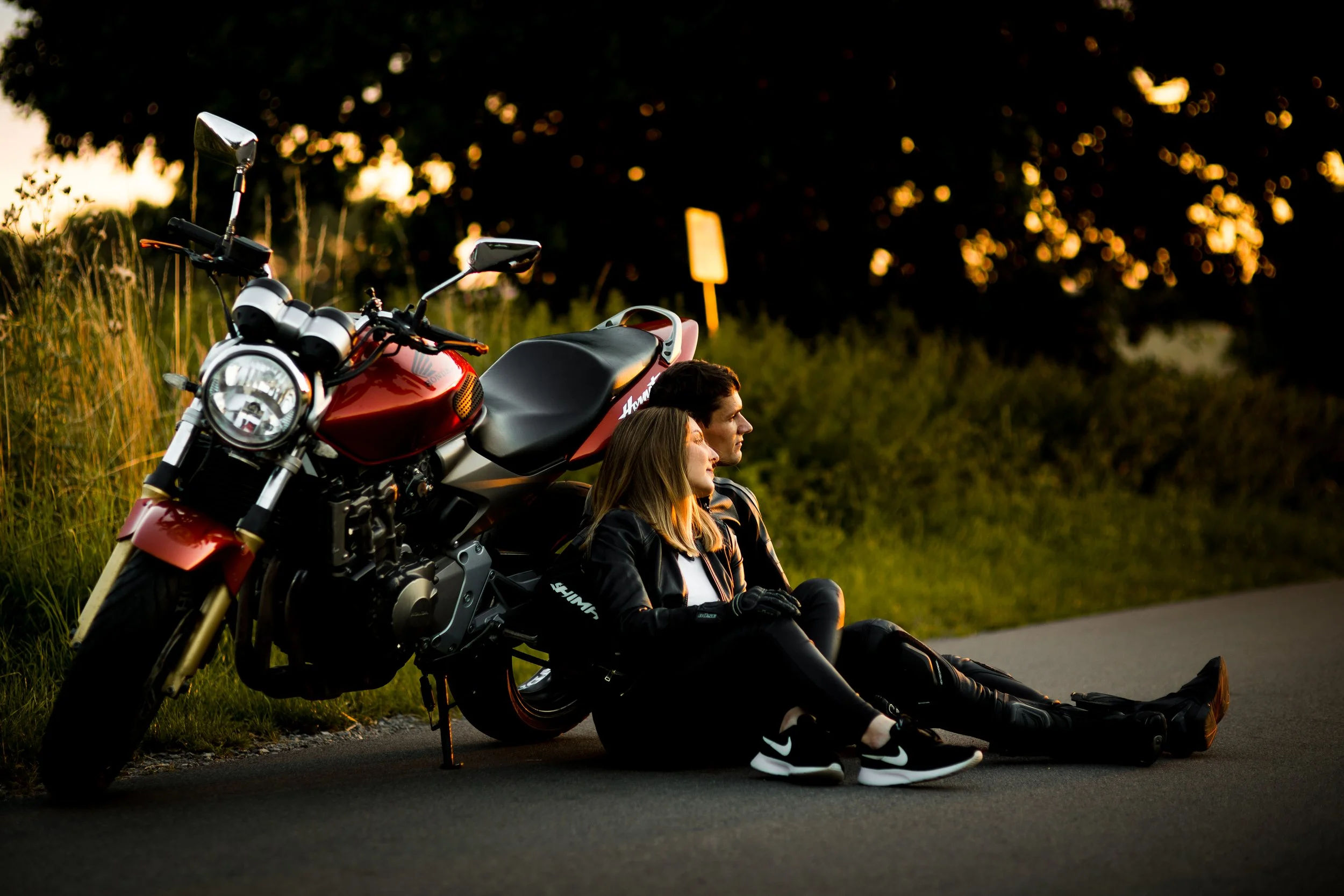A young man and woman sitting on the side of a road next to a red motorcycle, at sunset or dusk, with greenery and trees in the background.
