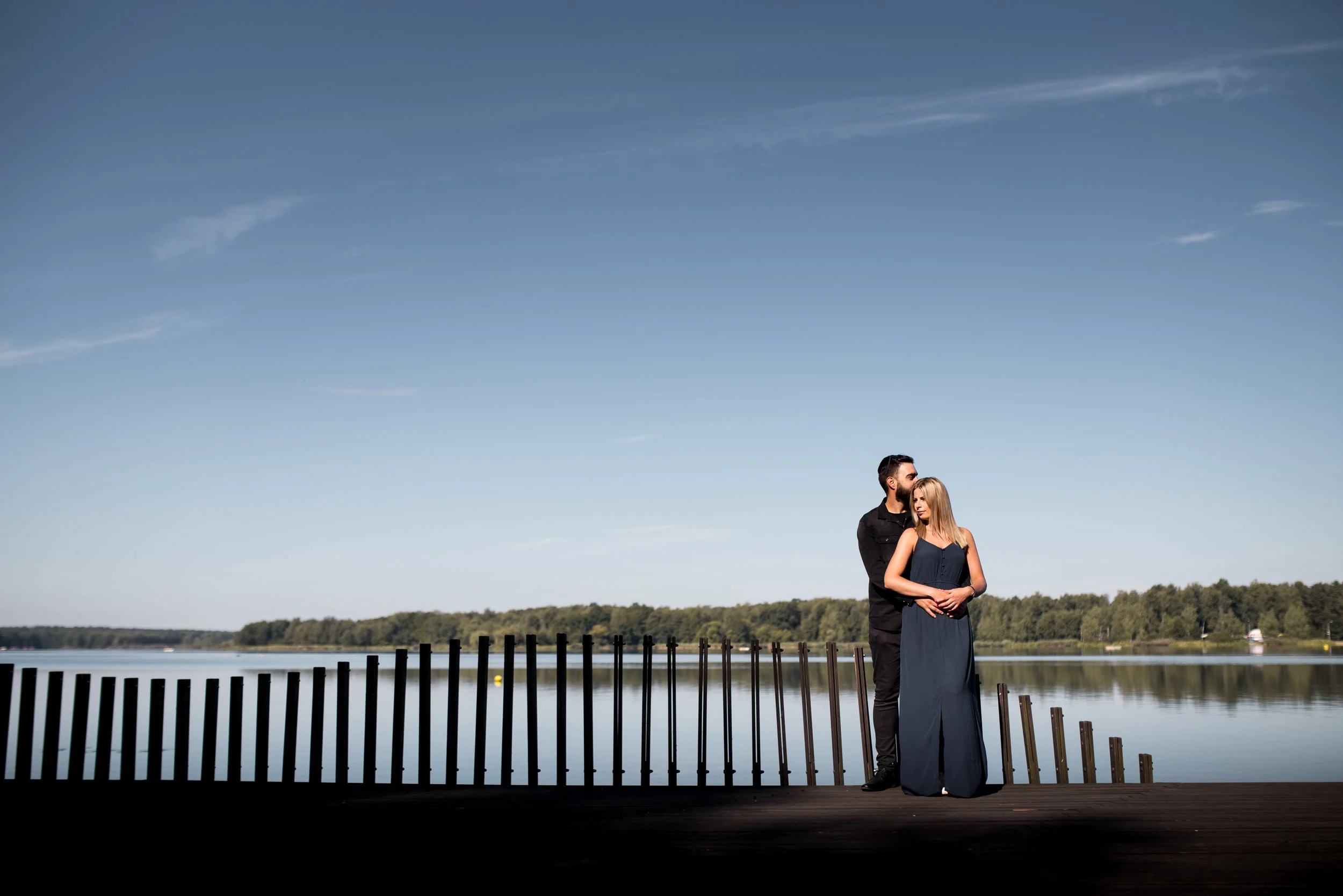 A couple standing on a wooden dock by a calm lake under a clear blue sky, with trees in the background.