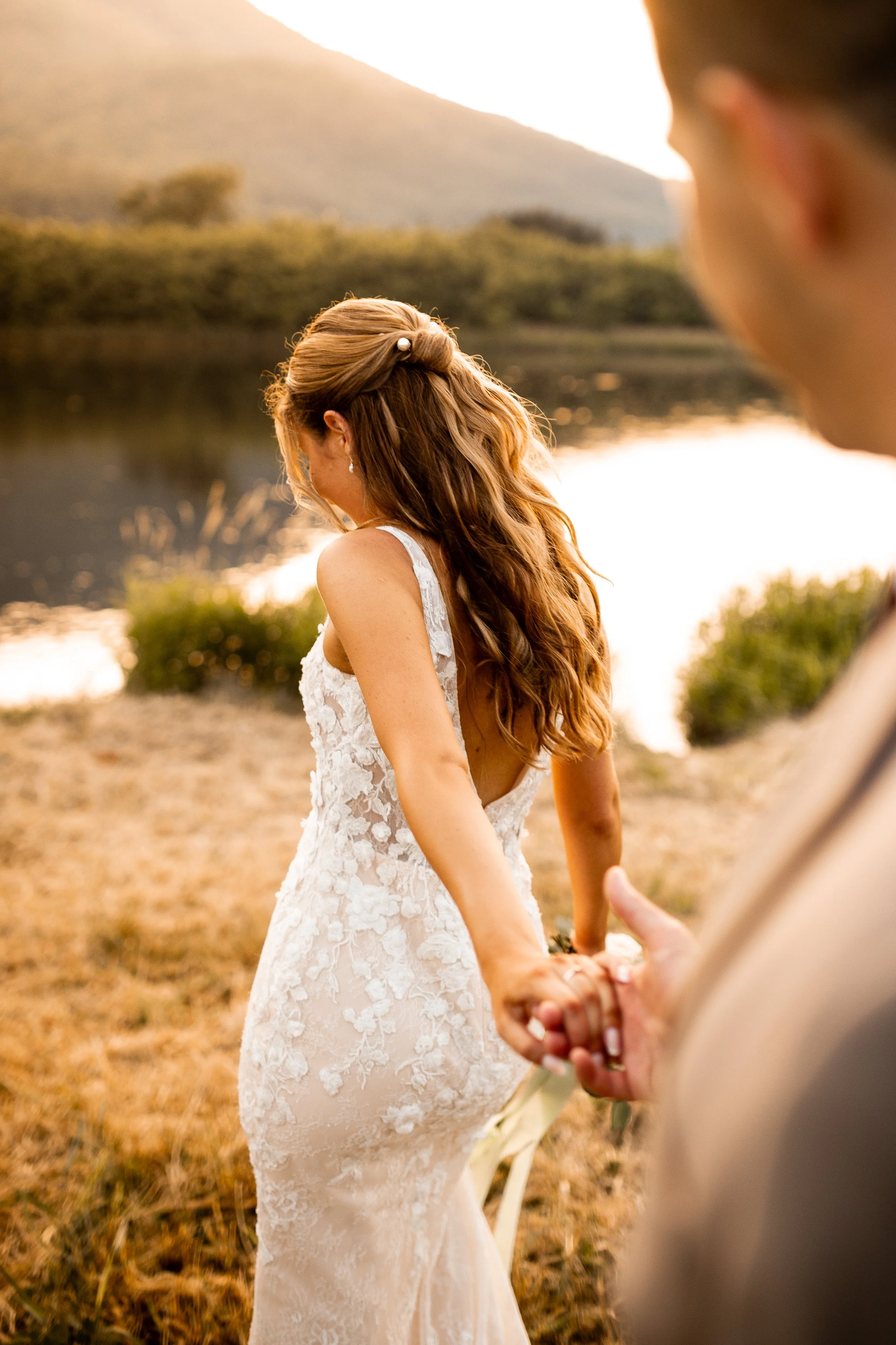 A bride in a lace wedding dress holding hands with her partner near a lakeside at sunset, with mountains in the background.