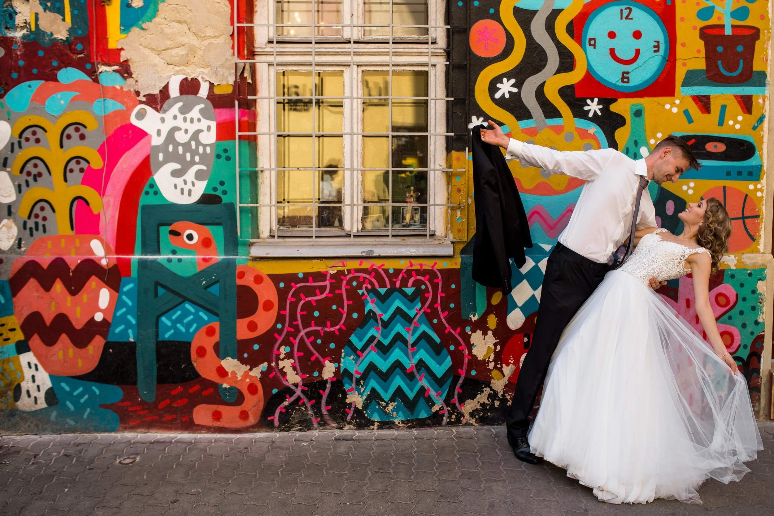 Para nowożeńców tańczy przed kolorową ścianą z graffiti, a pan młody zanurza pannę młodą w wodzie. Panna młoda ma na sobie białą suknię ślubną, a pan młody białą koszulę i czarny krawat.
