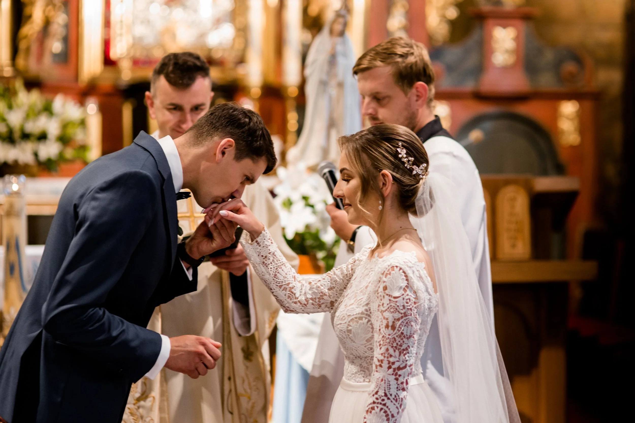 Pan młody całuje dłoń panny młodej podczas ceremonii ślubnej w kościele. Obecni są ksiądz i drużba.