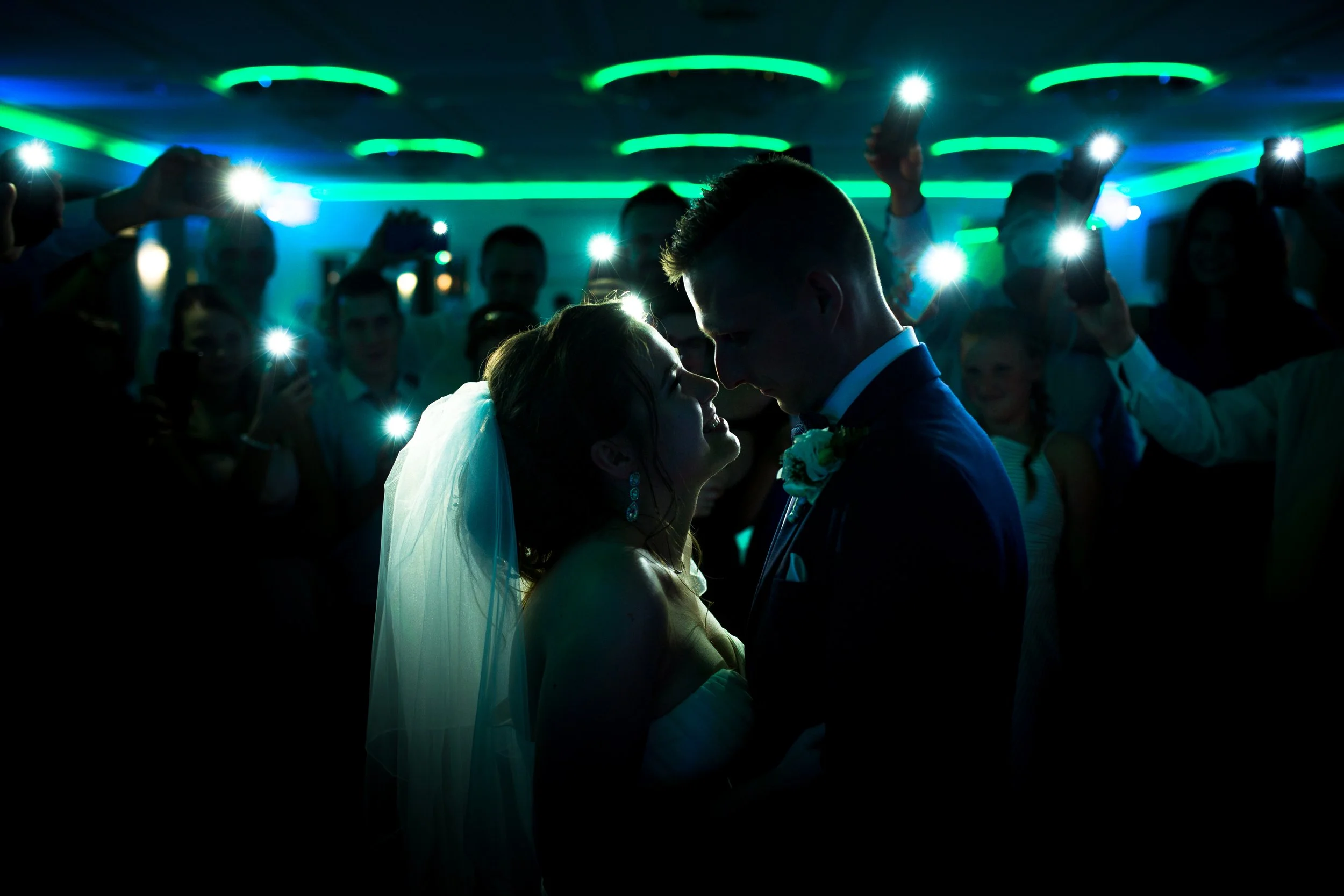 Para młoda tańczy na przyjęciu weselnym, w otoczeniu gości trzymających w rękach telefony komórkowe z włączonymi latarkami, w ciemnym pomieszczeniu z zielonymi i niebieskimi światłami na suficie.