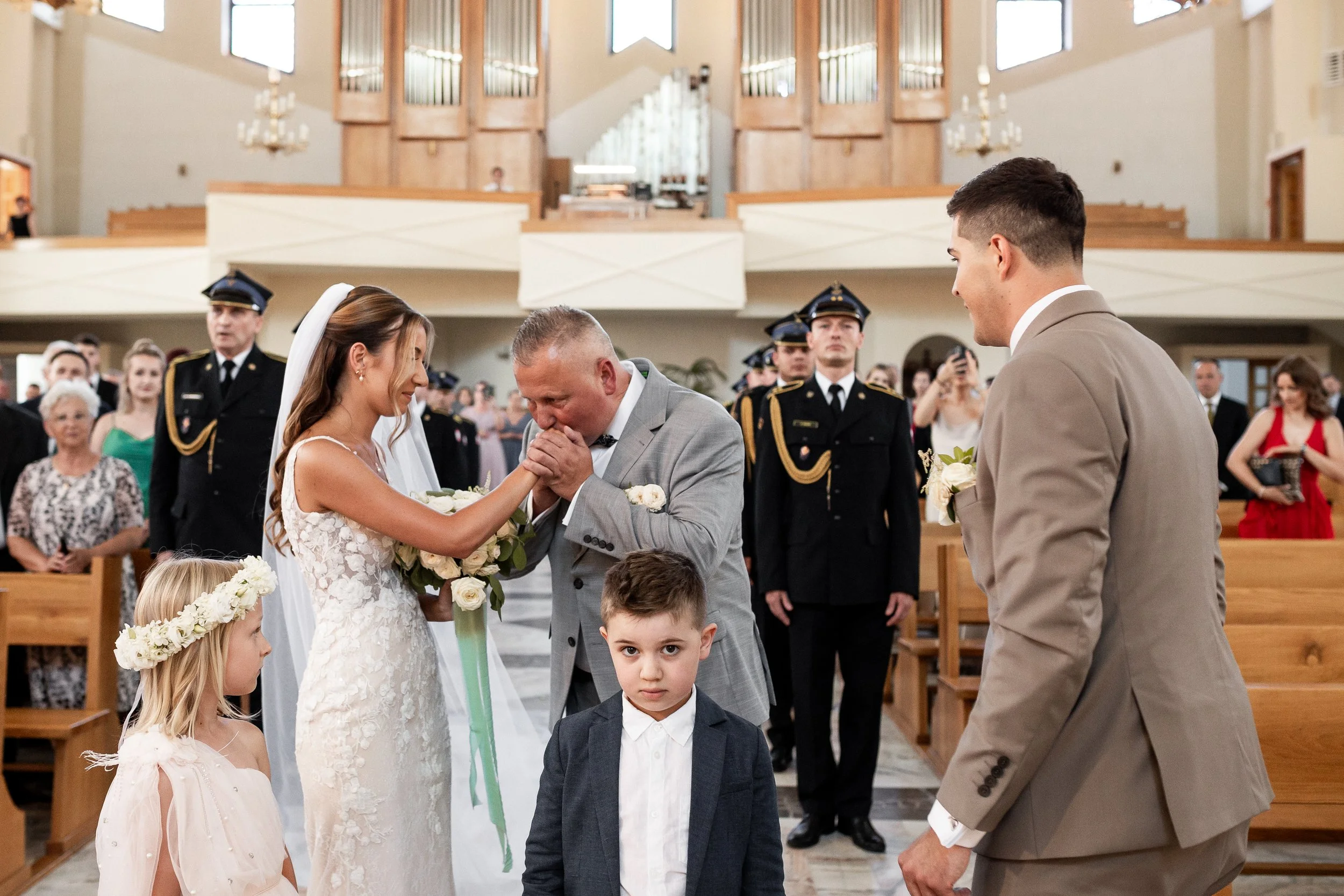 A wedding ceremony inside a church with a bride, groom, and a man, possibly the bride's father, holding hands and kissing. Two children, a girl with a flower crown and a boy, stand in the foreground. Guests and military personnel are visible in the b