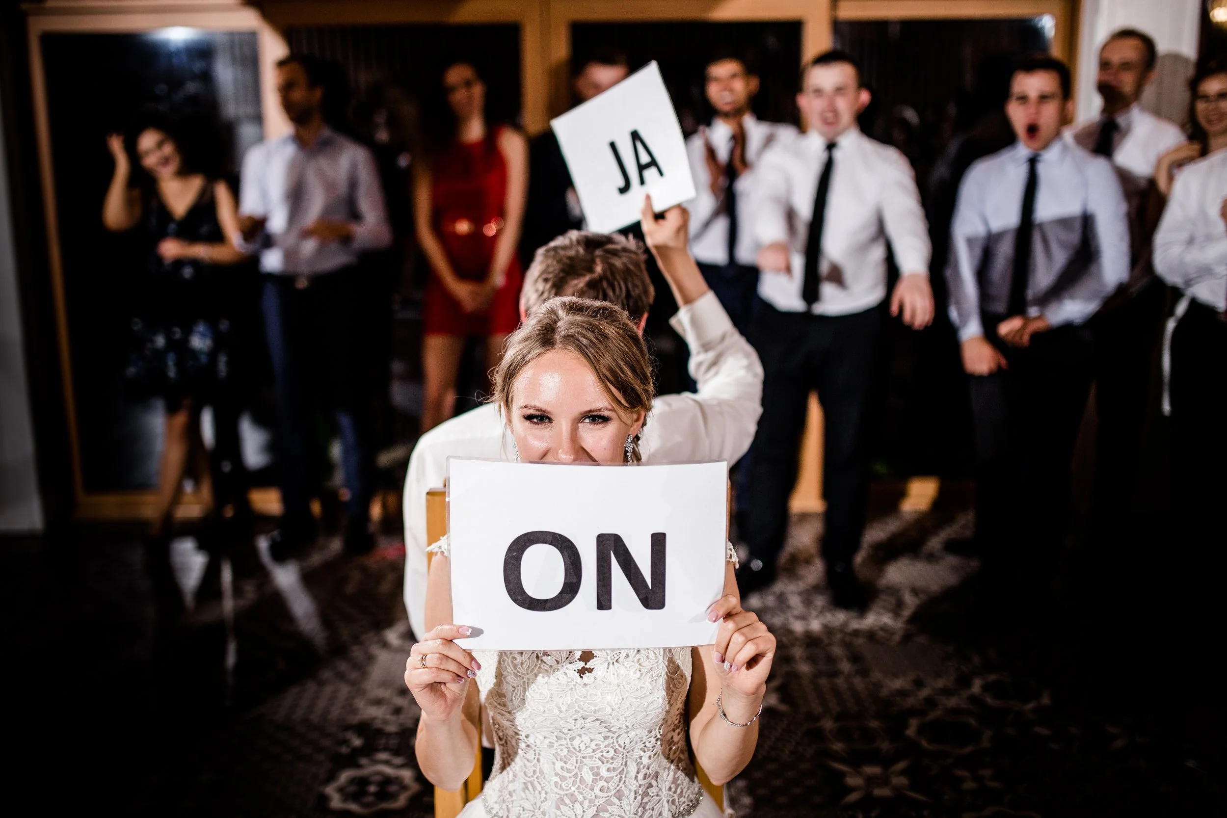 A smiling woman in a wedding dress holds a sign with 'ON' in front of her face. Behind her, a man in a white shirt and tie holds a sign with 'JA.' In the background, a group of people dressed in formal attire react with cheers and excitement.