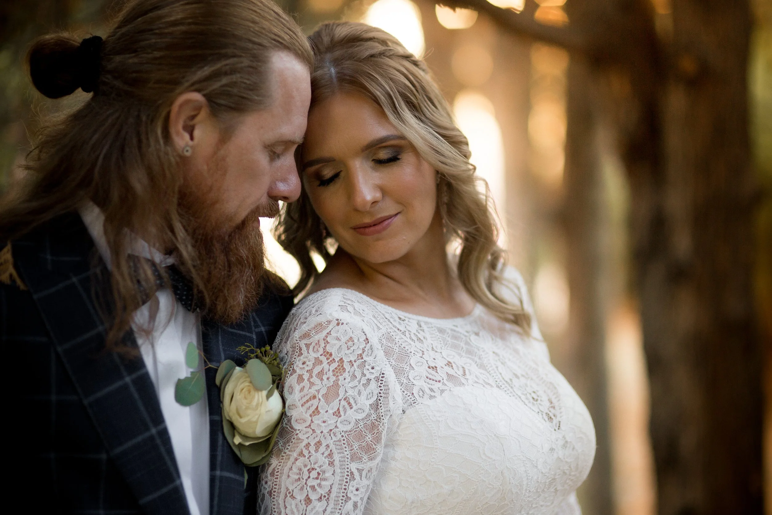 A man and woman are close together, touching foreheads with eyes closed, in a tender moment outdoors. The man has long hair tied back, a beard, and is wearing a checkered suit with a boutonniere. The woman has blonde curly hair and is wearing a white