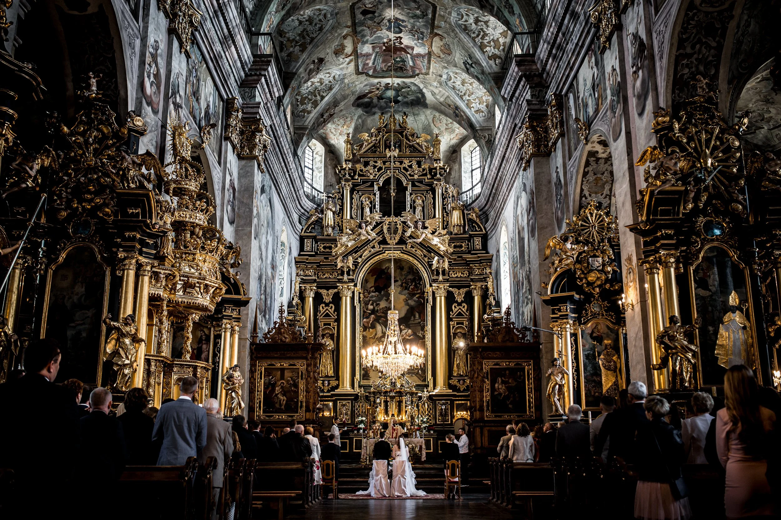 Interior of a richly decorated baroque church with gold accents, ornate statues, and a high vaulted ceiling, filled with people attending a ceremony.