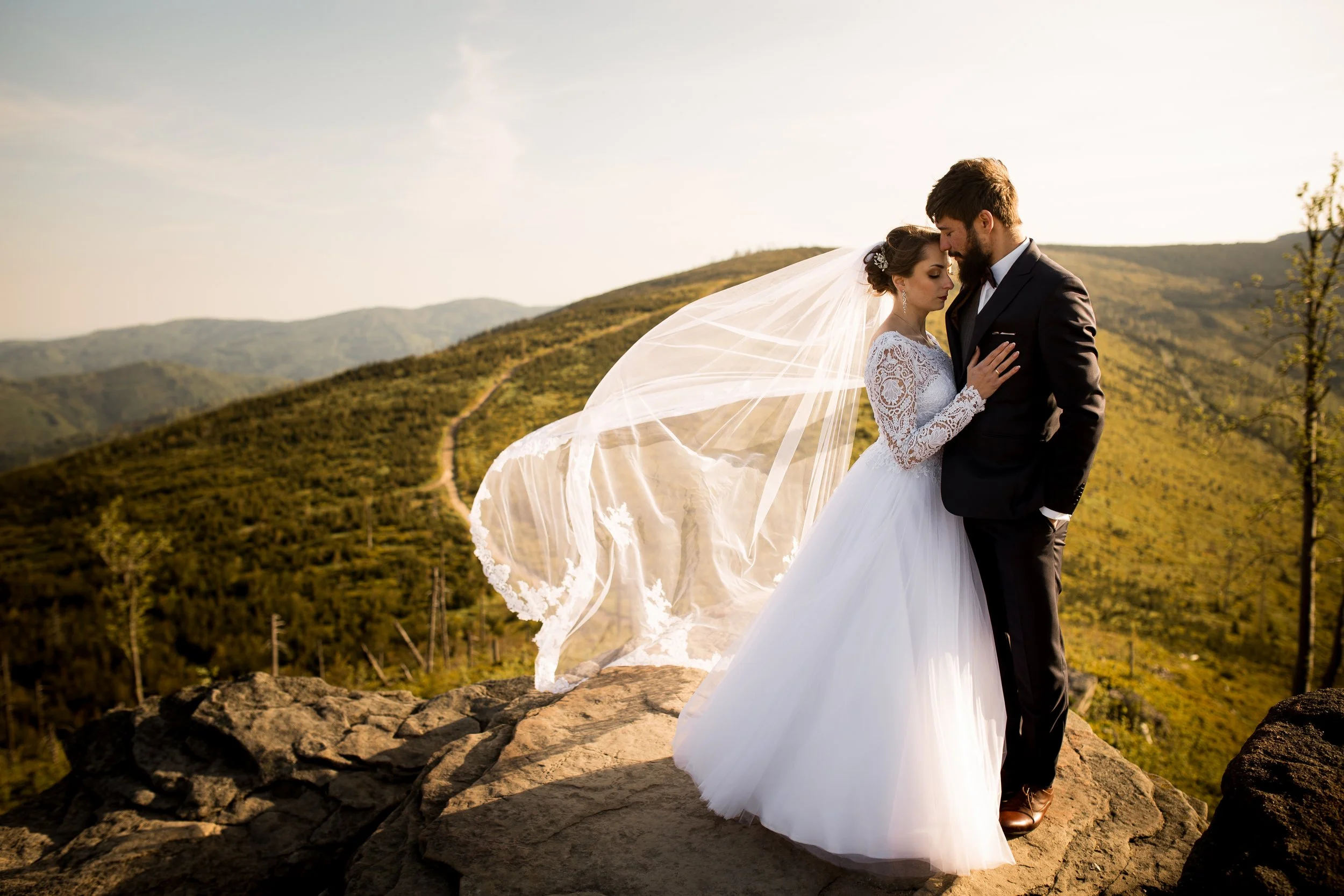 A bride and groom in wedding attire embracing on a rocky hilltop with a scenic mountain landscape in the background during sunset.