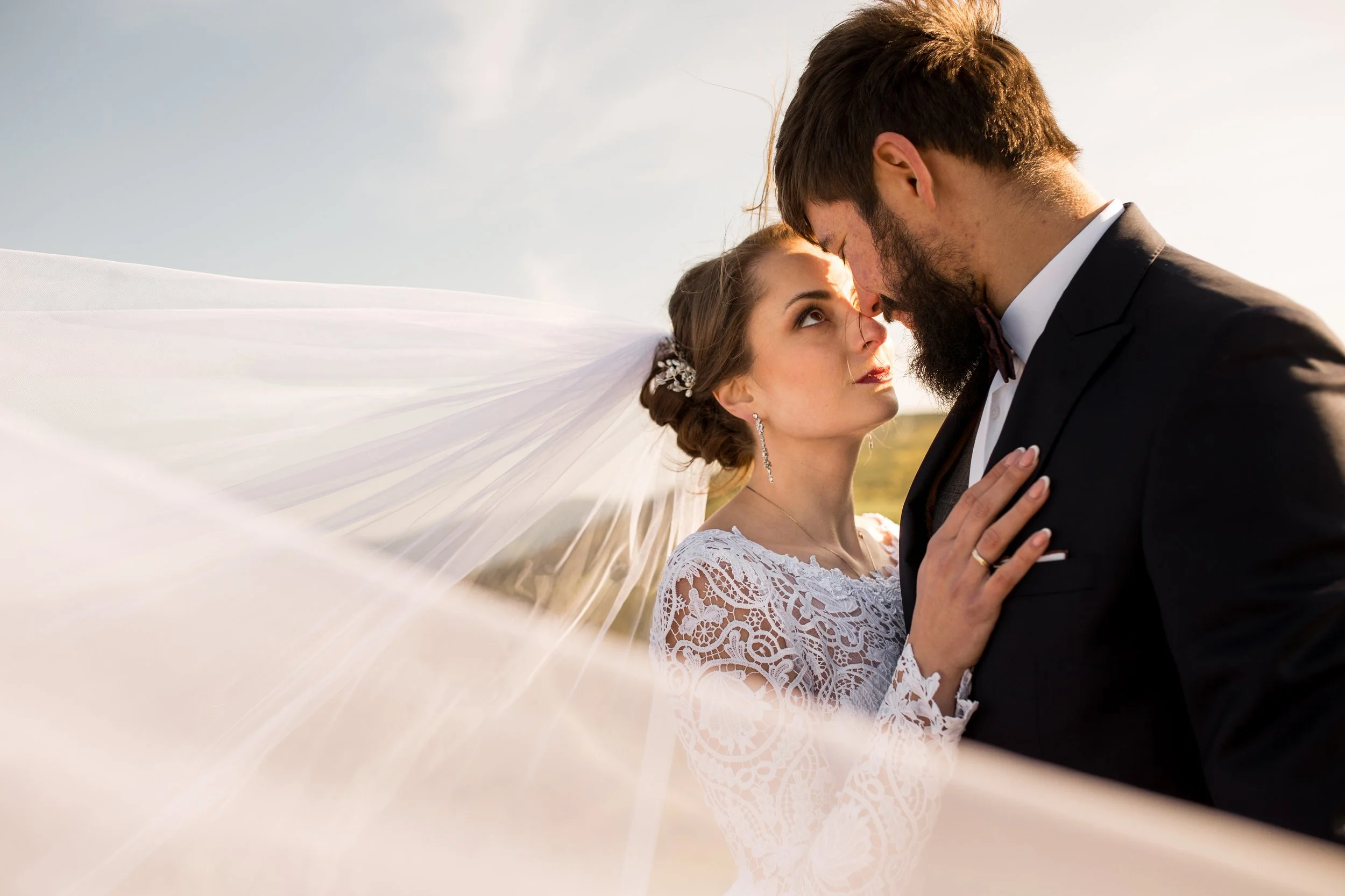 A bride and groom gently touching foreheads during a wedding photoshoot outdoors at sunset, with distant fields and a partially cloudy sky.